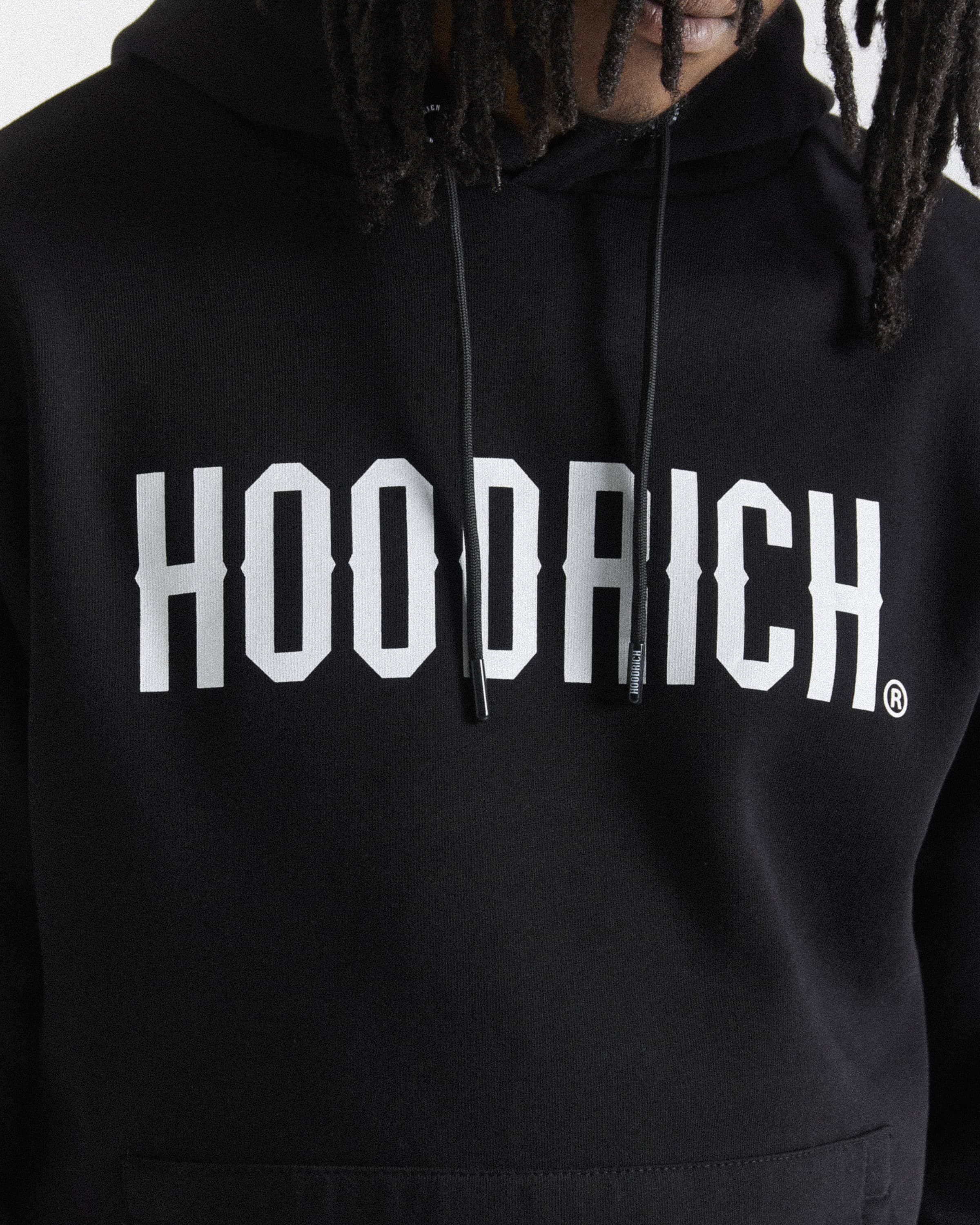 A close-up of a person wearing a black pullover hoodie with a large white 'HOODRICH' logo printed across the chest.
