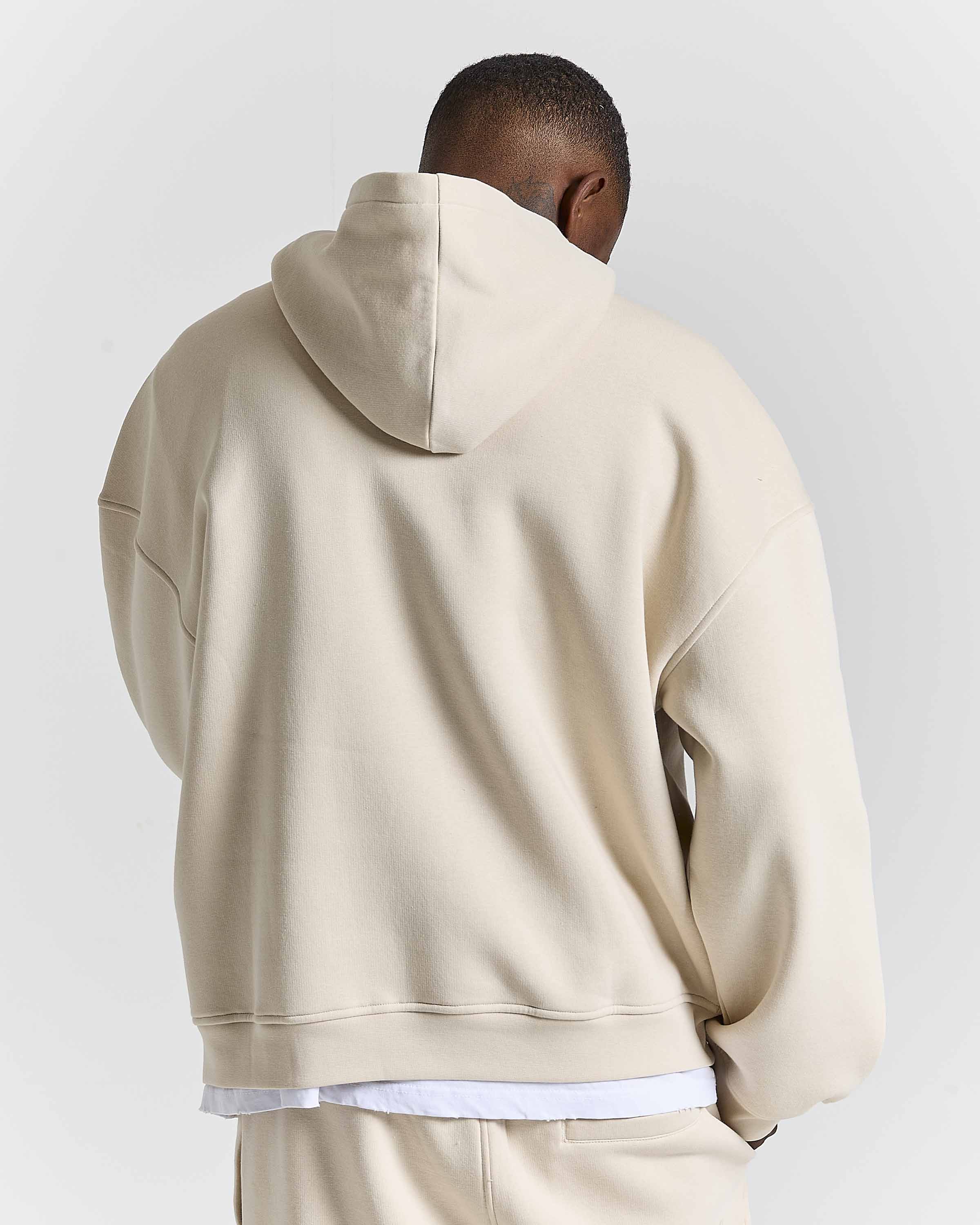 A man viewed from behind wearing a light beige oversized hoodie and matching sweatpants against a plain light gray studio background.