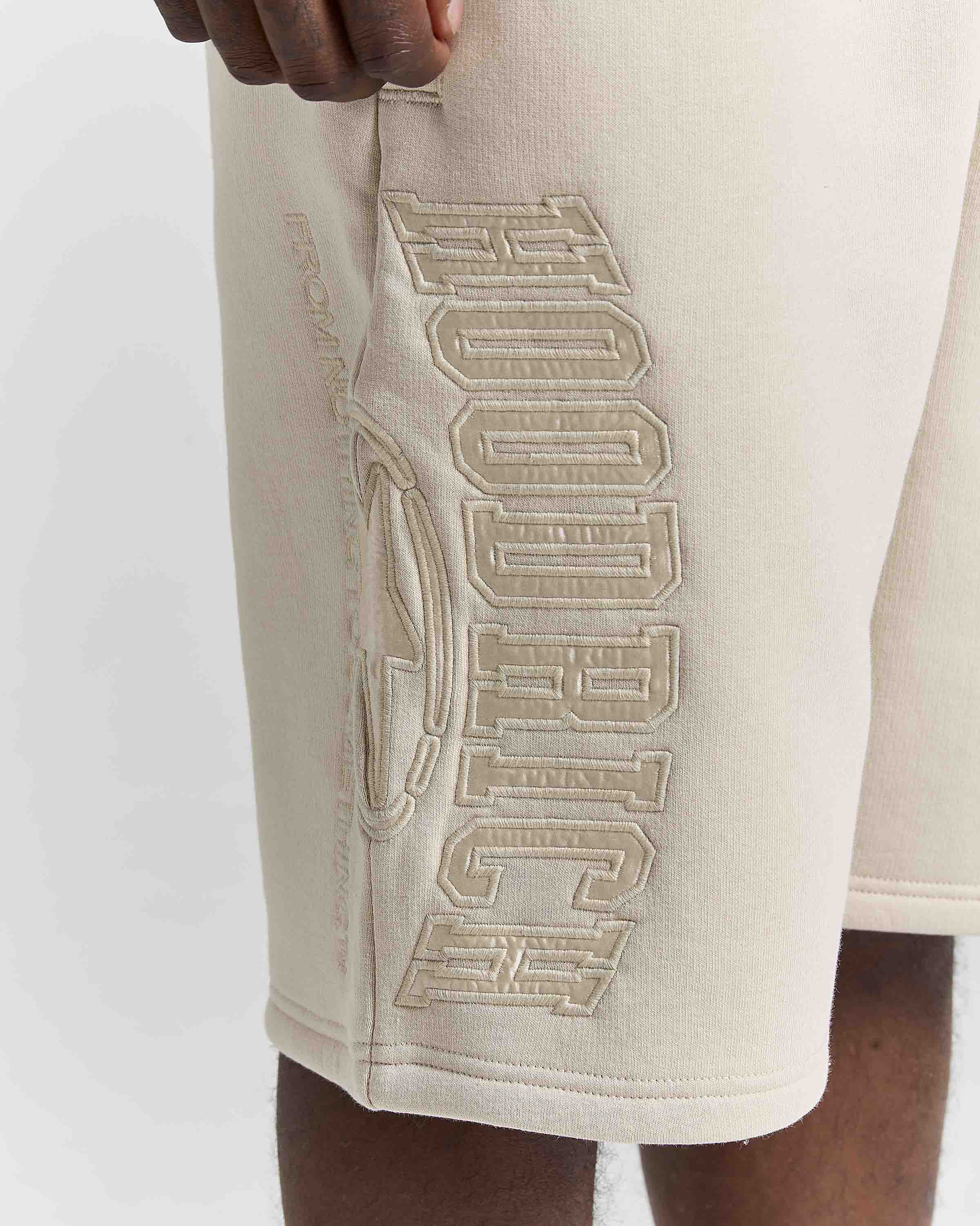 Close-up of a person wearing cream-colored athletic shorts with large tonal embroidered lettering on the side.