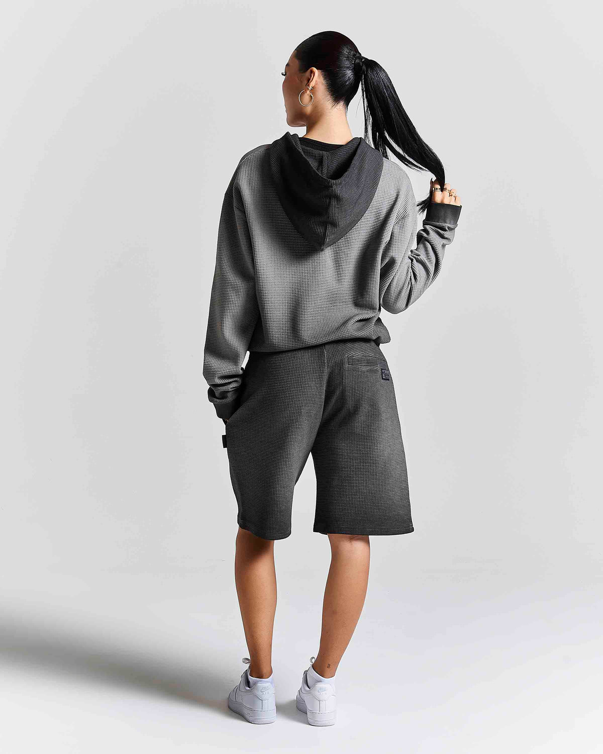 A woman shown from the back wearing a gray and black hooded sweatshirt, dark gray shorts, and white sneakers against a light gray studio background.
