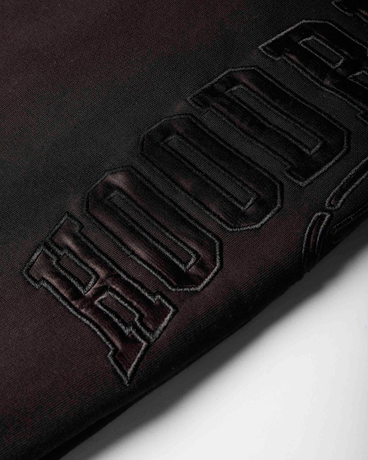 Close-up of a black fabric surface with large raised embroidered lettering in a dark metallic burgundy tone, photographed on a white background.