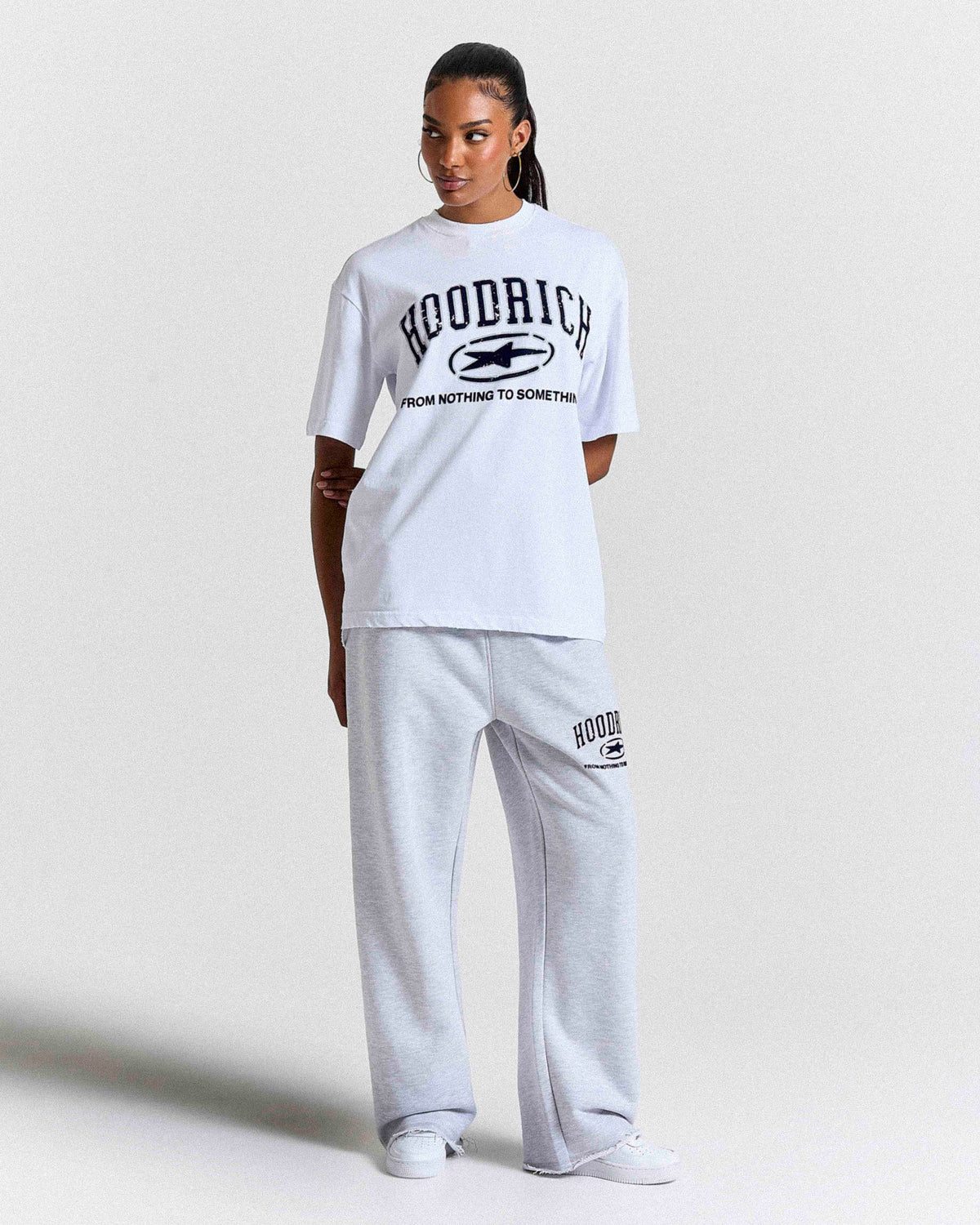 A model wearing a white short-sleeve graphic t-shirt and light gray sweatpants with black 'HOODRICH' branding, standing against a light gray studio background.