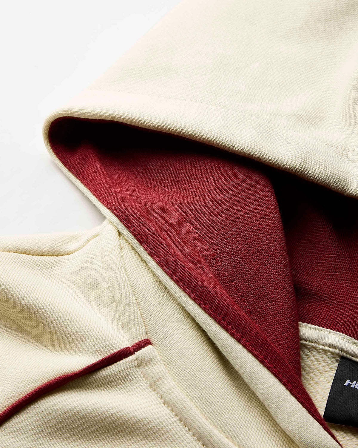 Close-up detail of a cream-colored hooded garment with a contrasting dark red inner lining and a small black brand tag.