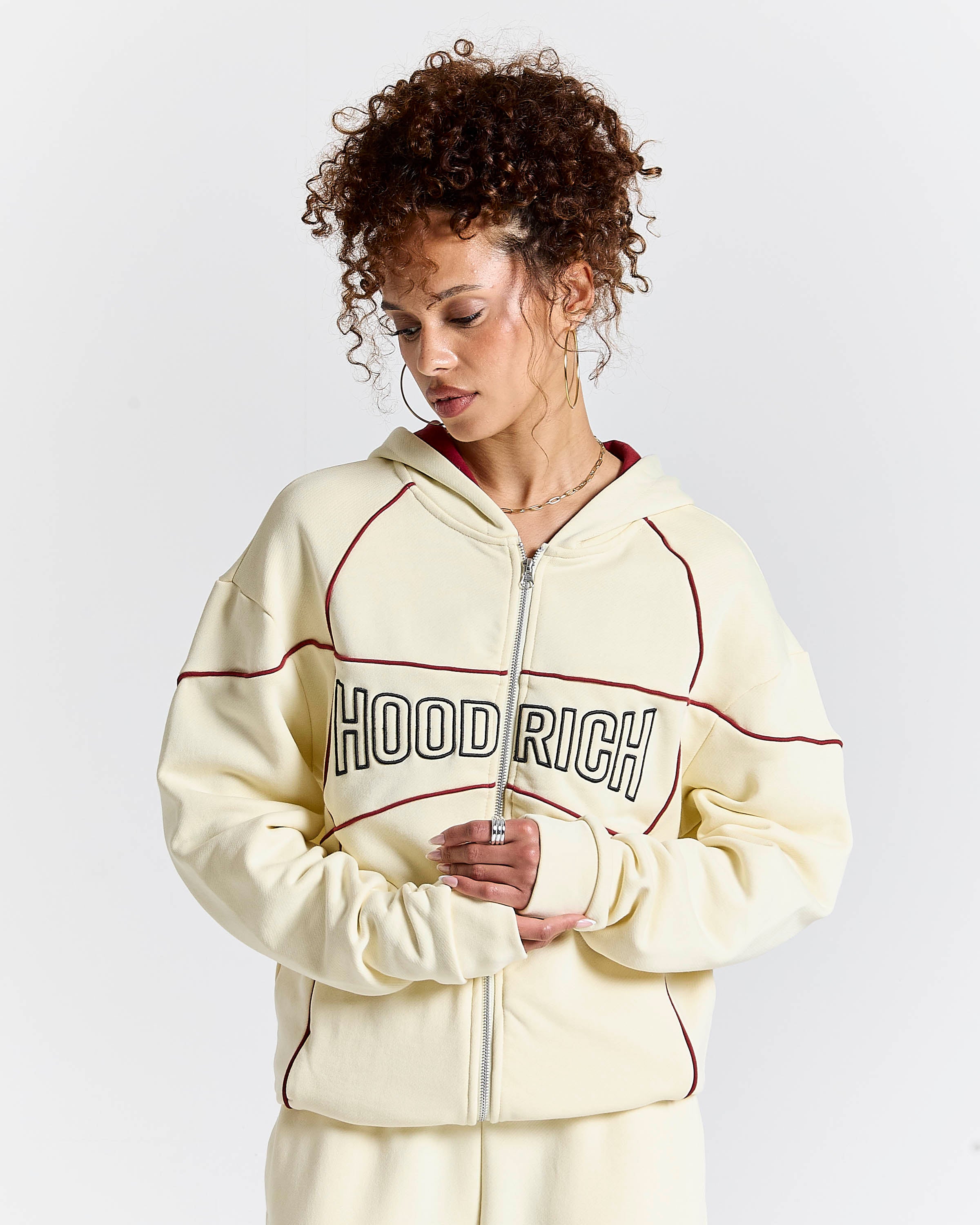 A woman wearing a cream zip-up hoodie with 'HOODRICH' printed across the chest in black outlined letters, accented with thin red piping, set against a plain light gray background.