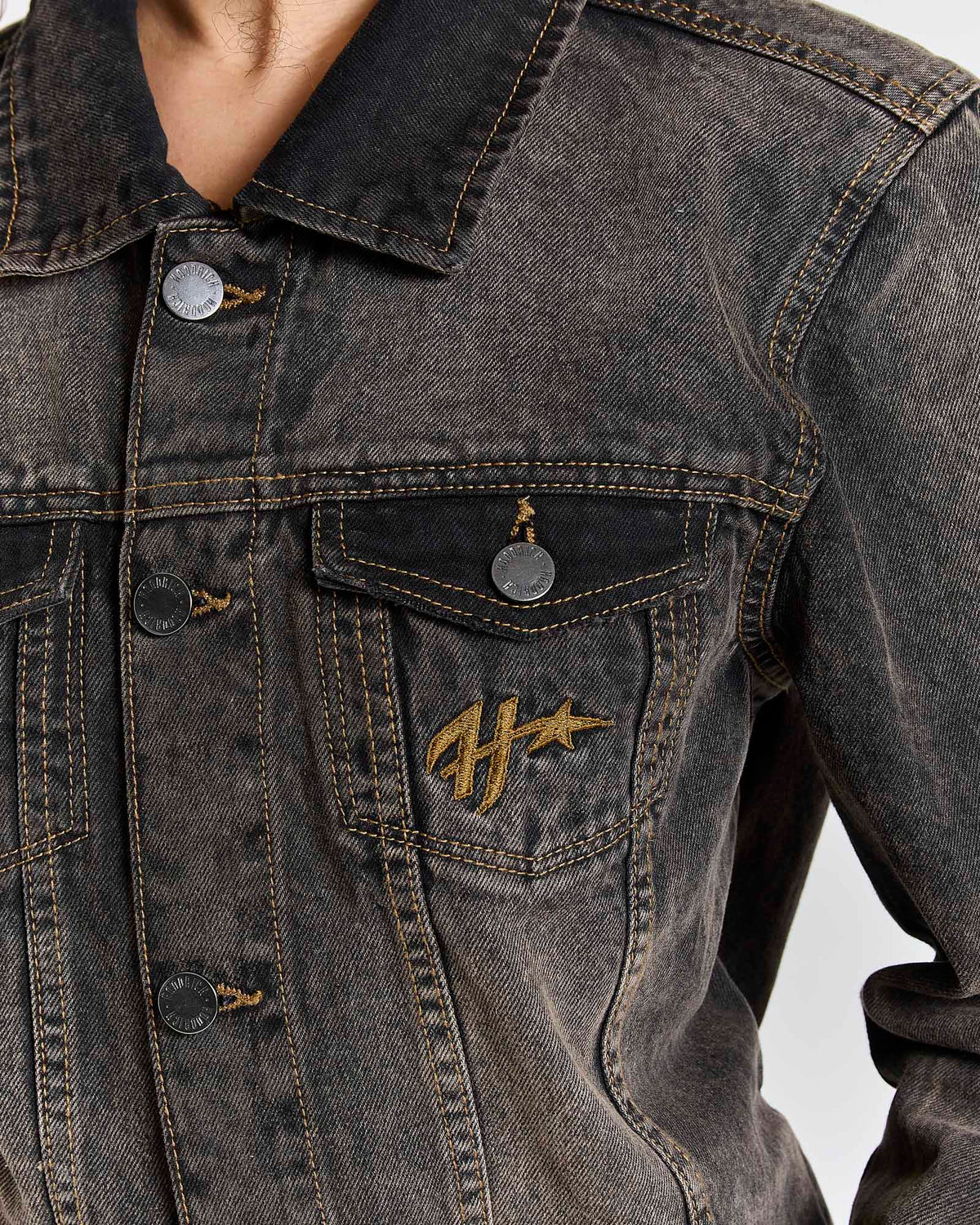 Close-up of a dark gray washed denim jacket with a gold embroidered logo on the chest pocket, featuring metal snap buttons and a collar.