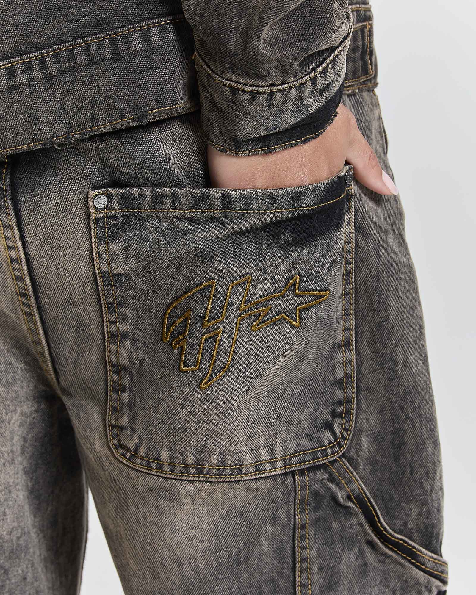 Close-up of a person wearing washed charcoal gray denim jeans with a gold embroidered Hoodrich logo on the back pocket, showing a hand in the pocket.