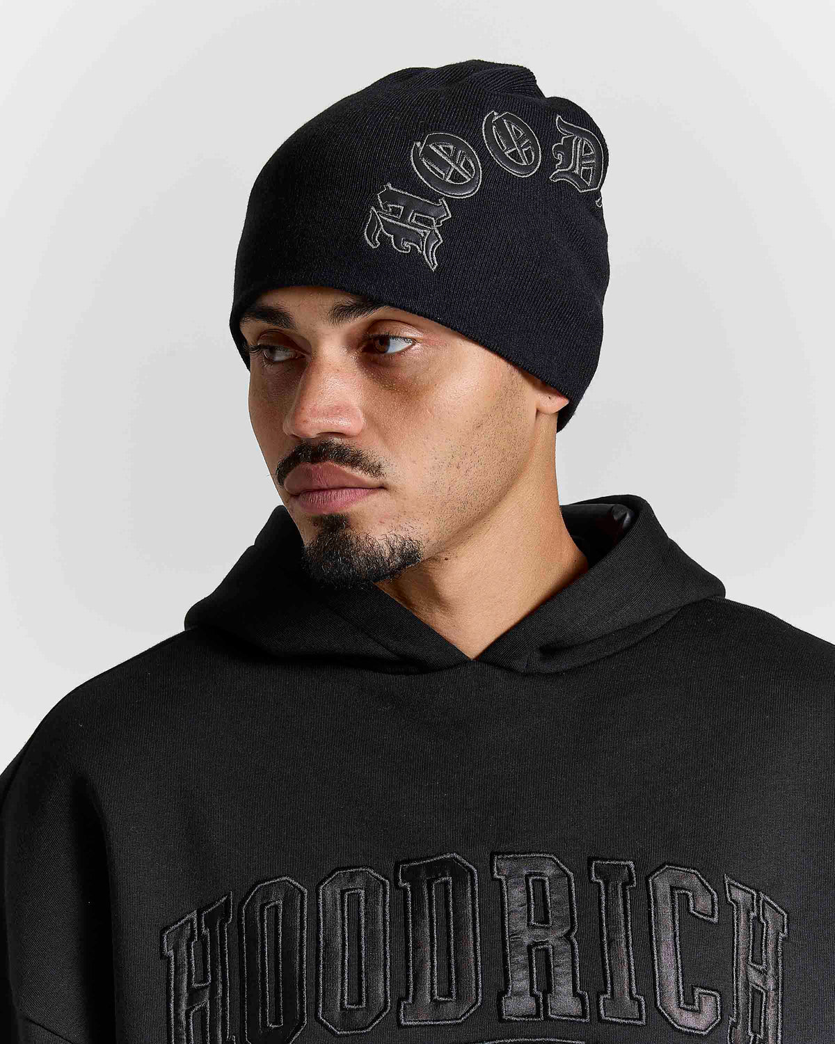 A man wearing a black knit beanie with a gray 'HOOD' graphic and a matching black hoodie with 'HOODRICH' lettering, photographed against a light gray studio background.