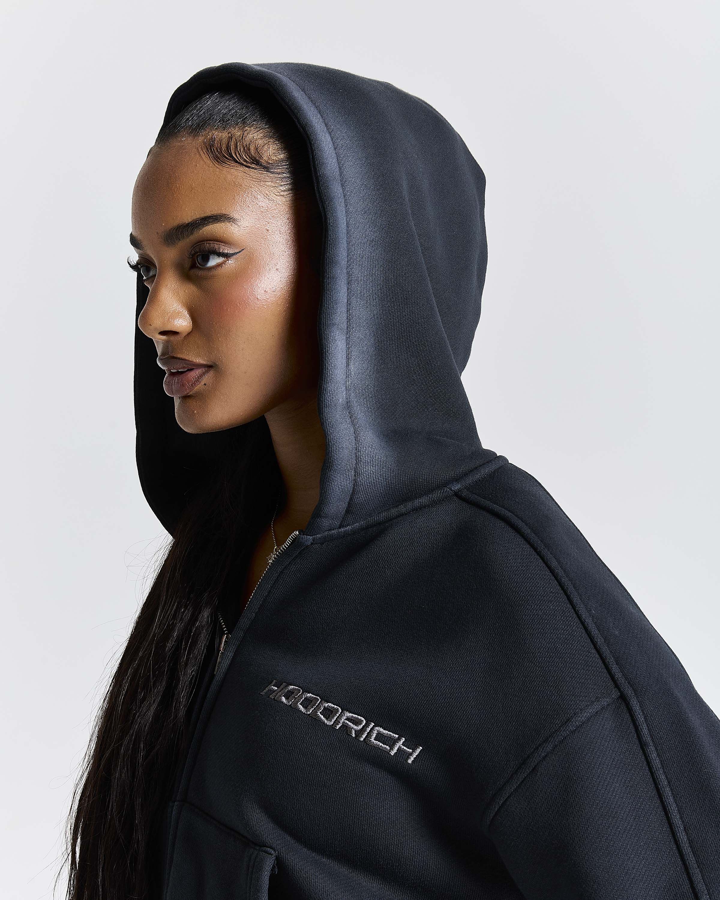 Close-up of a model wearing a black cropped zip-up hoodie, focusing on the hood and the Hoodrich chest logo.