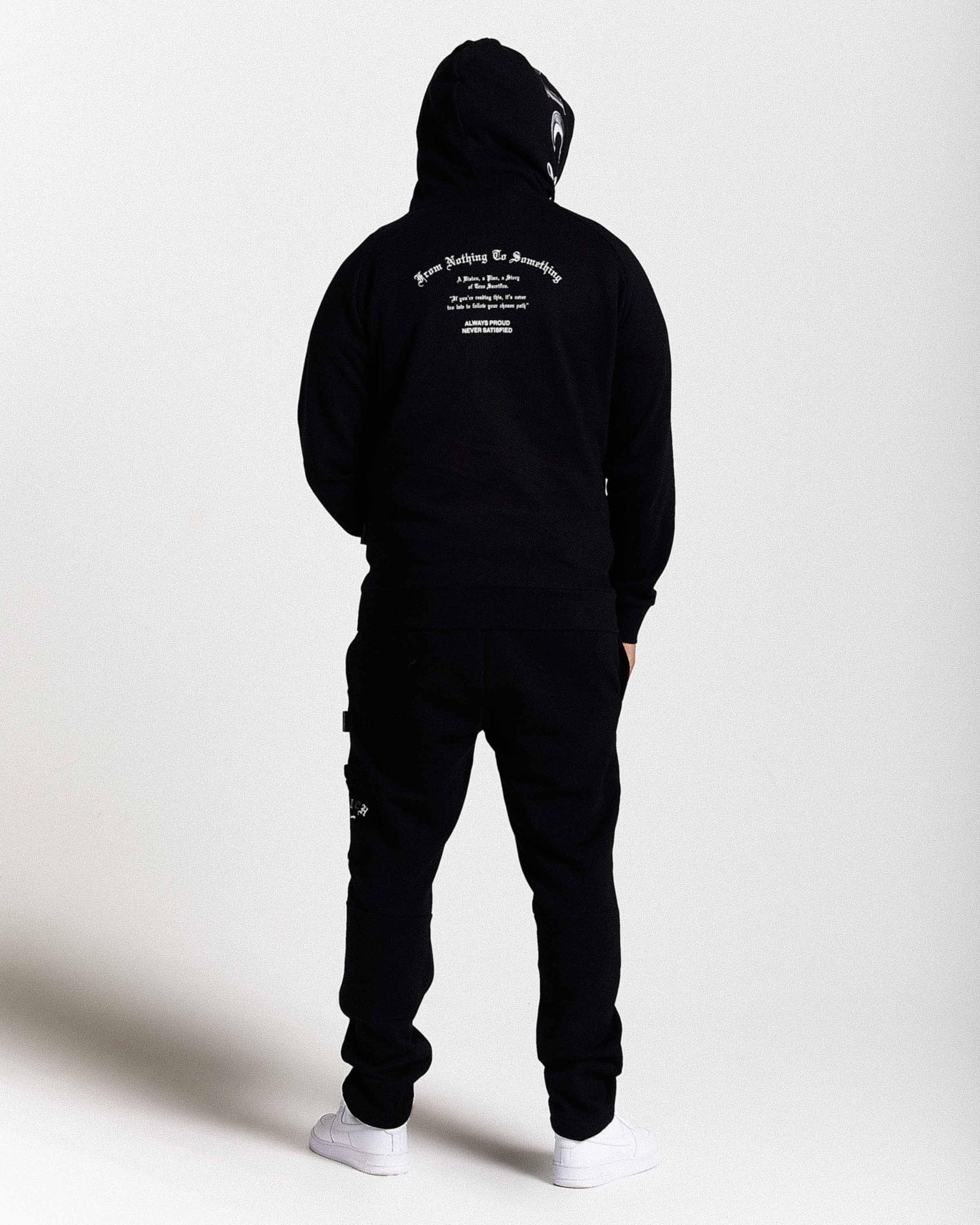 Displays a model wearing the Black 'Hoodrich' branded men's Stature Full Tracksuit