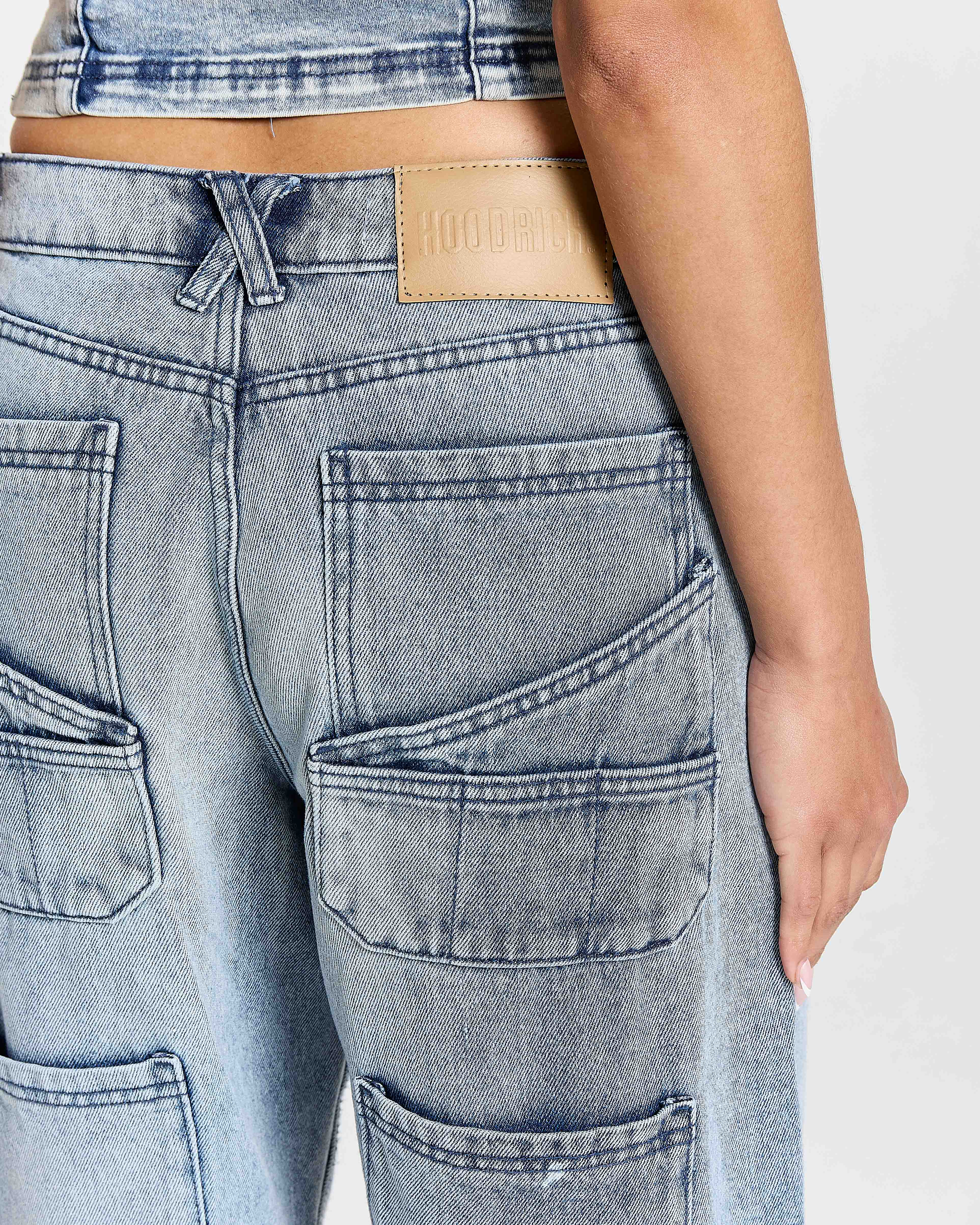 Close-up of a person wearing light blue jeans with a visible brand patch on a white background