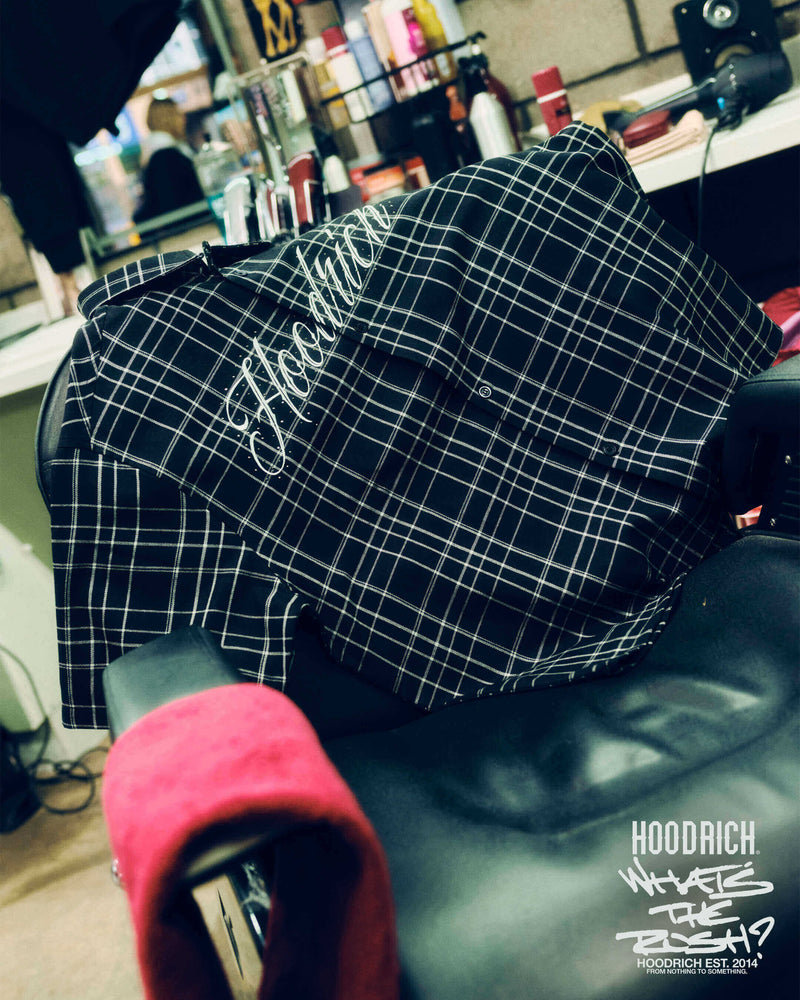 A black and white plaid hooded sweatshirt draped over a black salon chair in a barbershop or hair salon environment. The hoodie features a white script logo reading 'Hoodrich' on the chest.