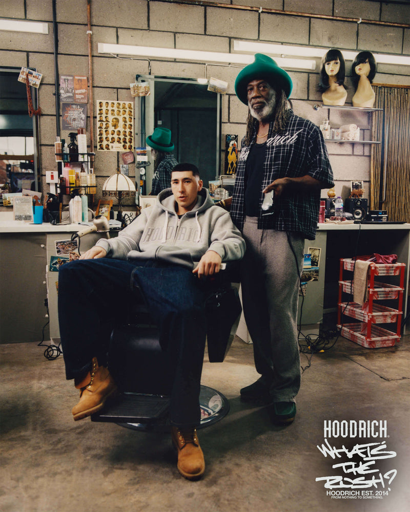 Two men in a barbershop with one sitting in a chair and the other standing, wearing a green hat and plaid shirt.