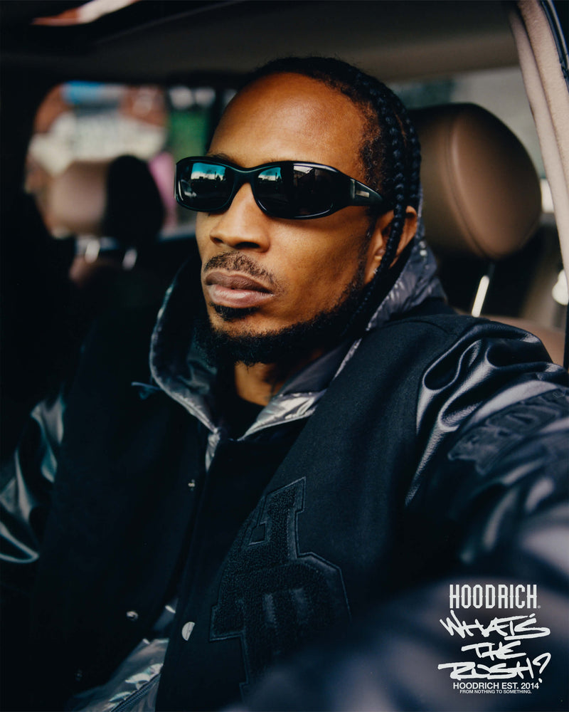 Man wearing sunglasses and a black jacket sitting inside a car, with 'Hoodrich' branding visible.