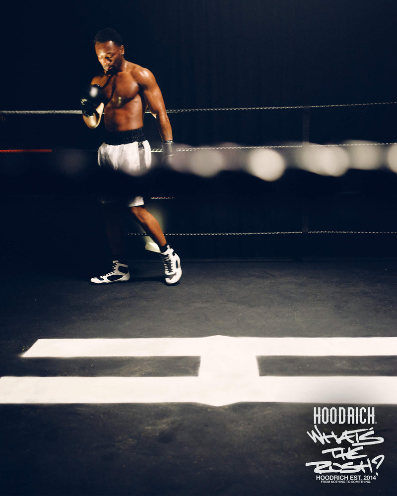 A shirtless male boxer in a ring wearing white and black shorts, black gloves, and white sneakers, posed in a dark boxing arena with a blurred rope in the foreground and a text overlay in the bottom right corner.