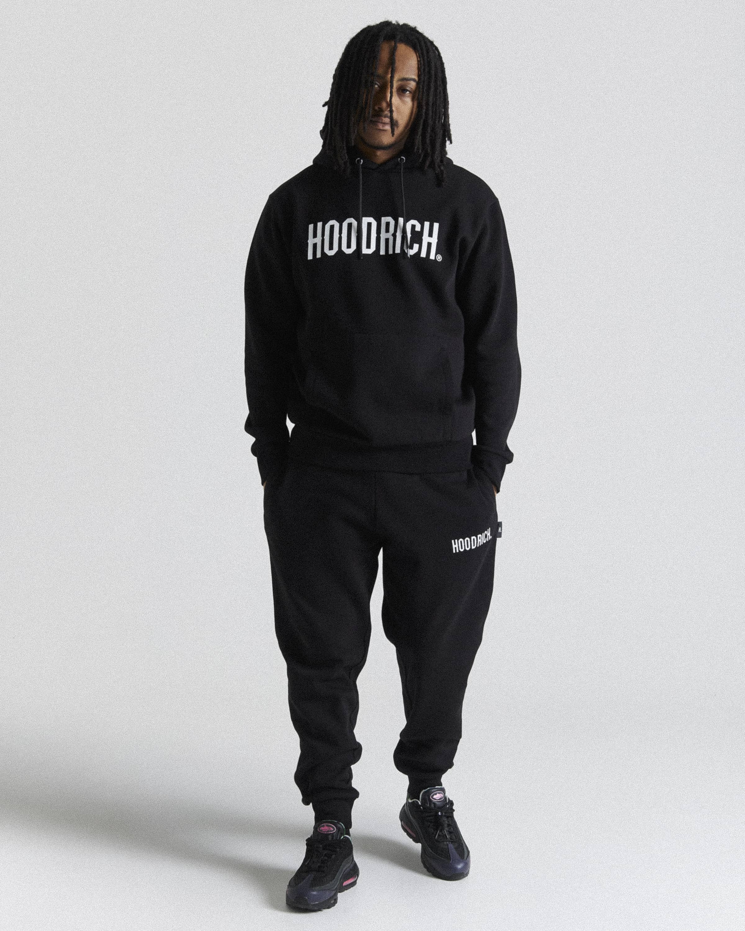 A man modeling a black hoodie and sweatpants set with white 'HOODRICH' branding, standing against a light gray studio background.