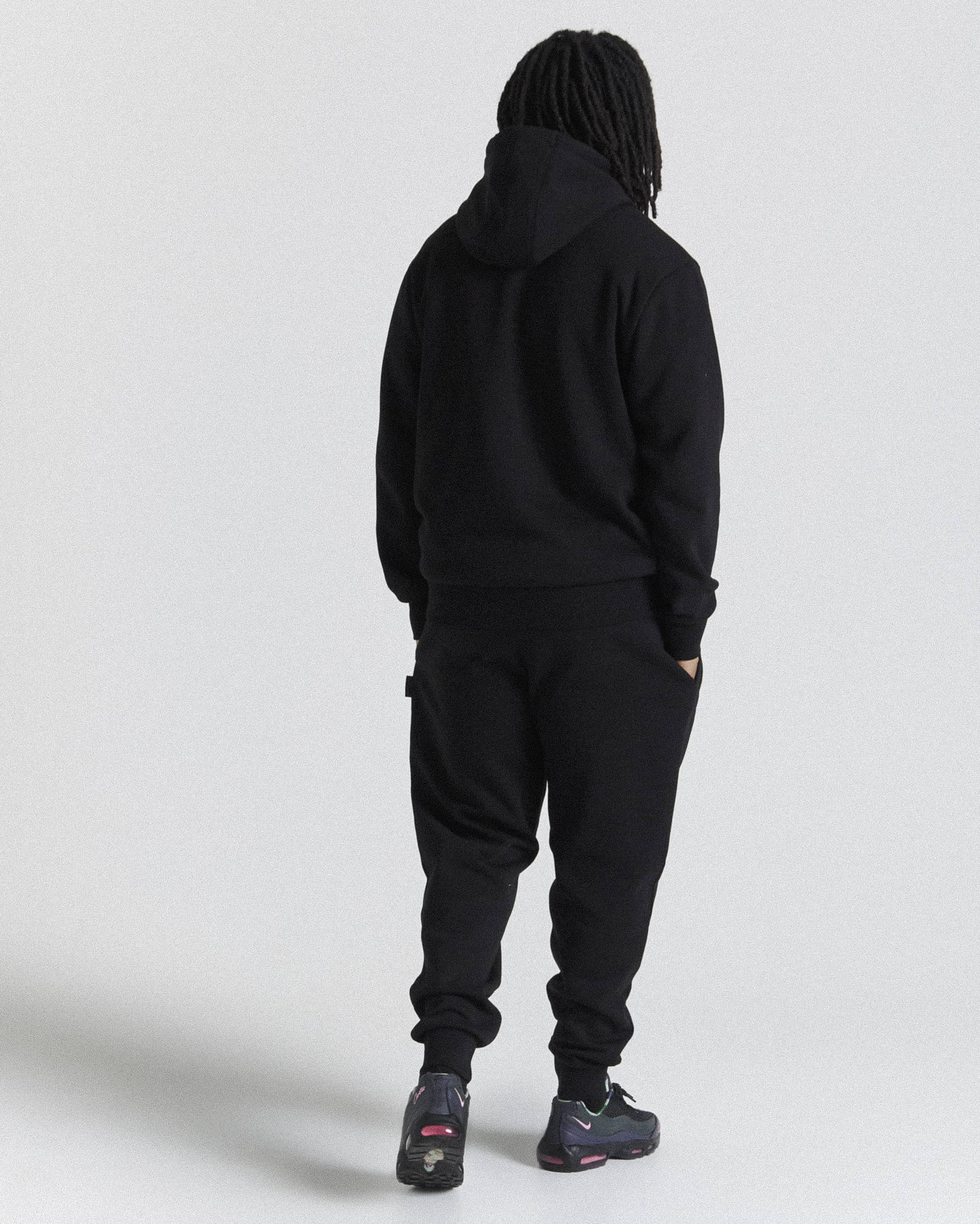 A person shown from the back wearing a black hooded sweatshirt and black jogger pants against a light gray studio background.