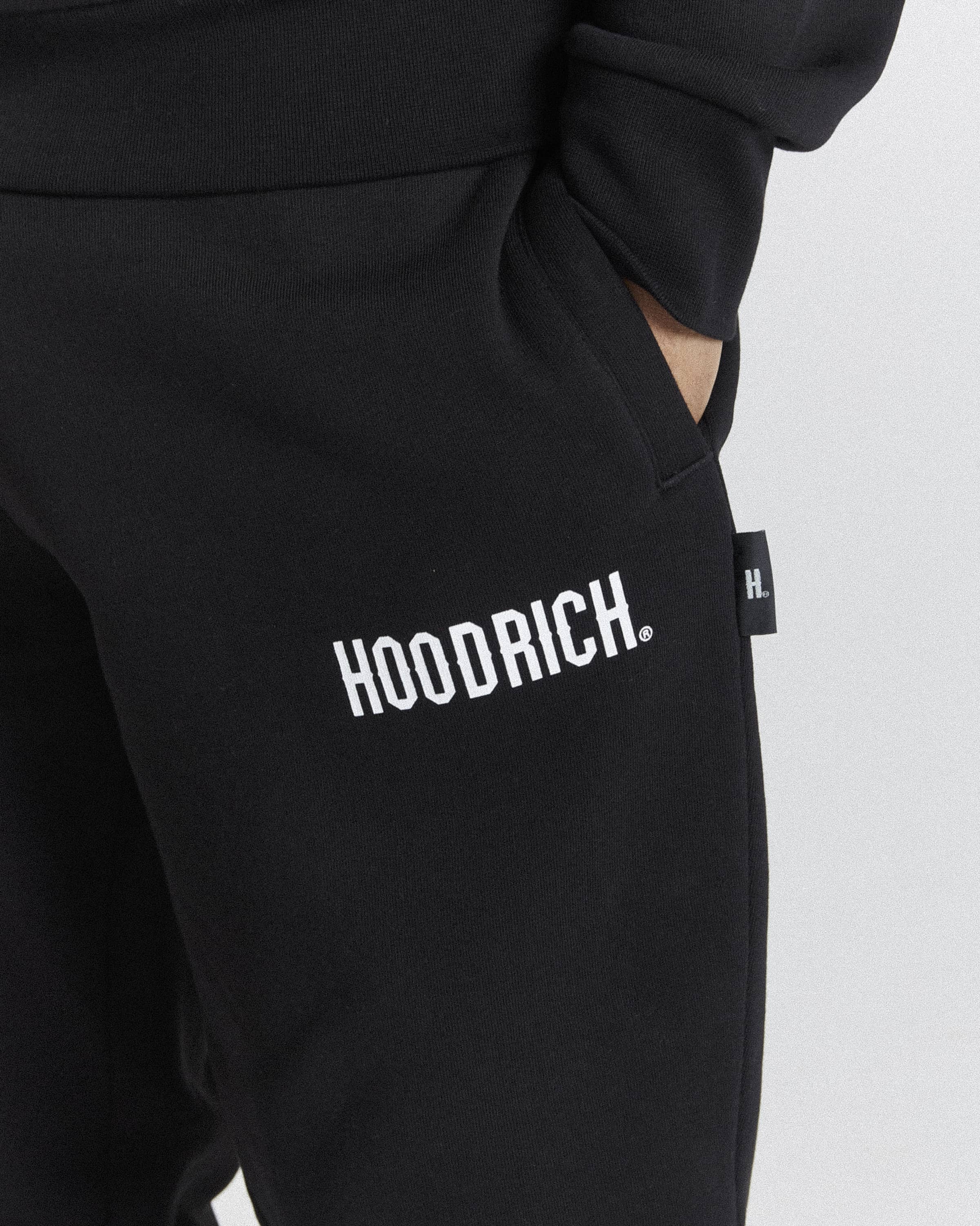 Close-up of black sweatpants with a white 'HOODRICH.' logo printed on the thigh and a small black side tag, shown on a model against a light gray background.
