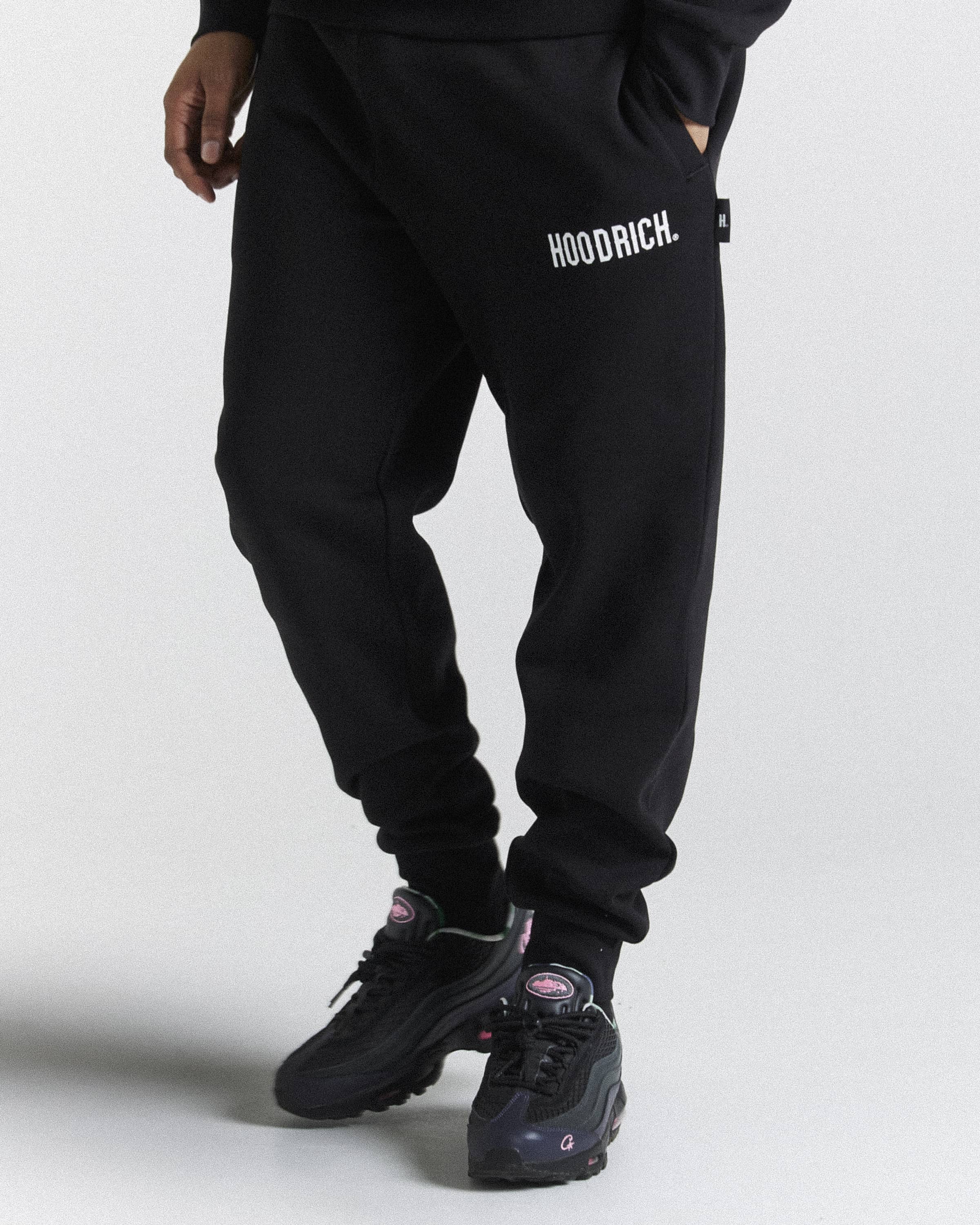 Black sweatpants with white 'HOODRICH.' text on the left thigh, shown on a model from the waist down wearing black sneakers against a light gray studio background.