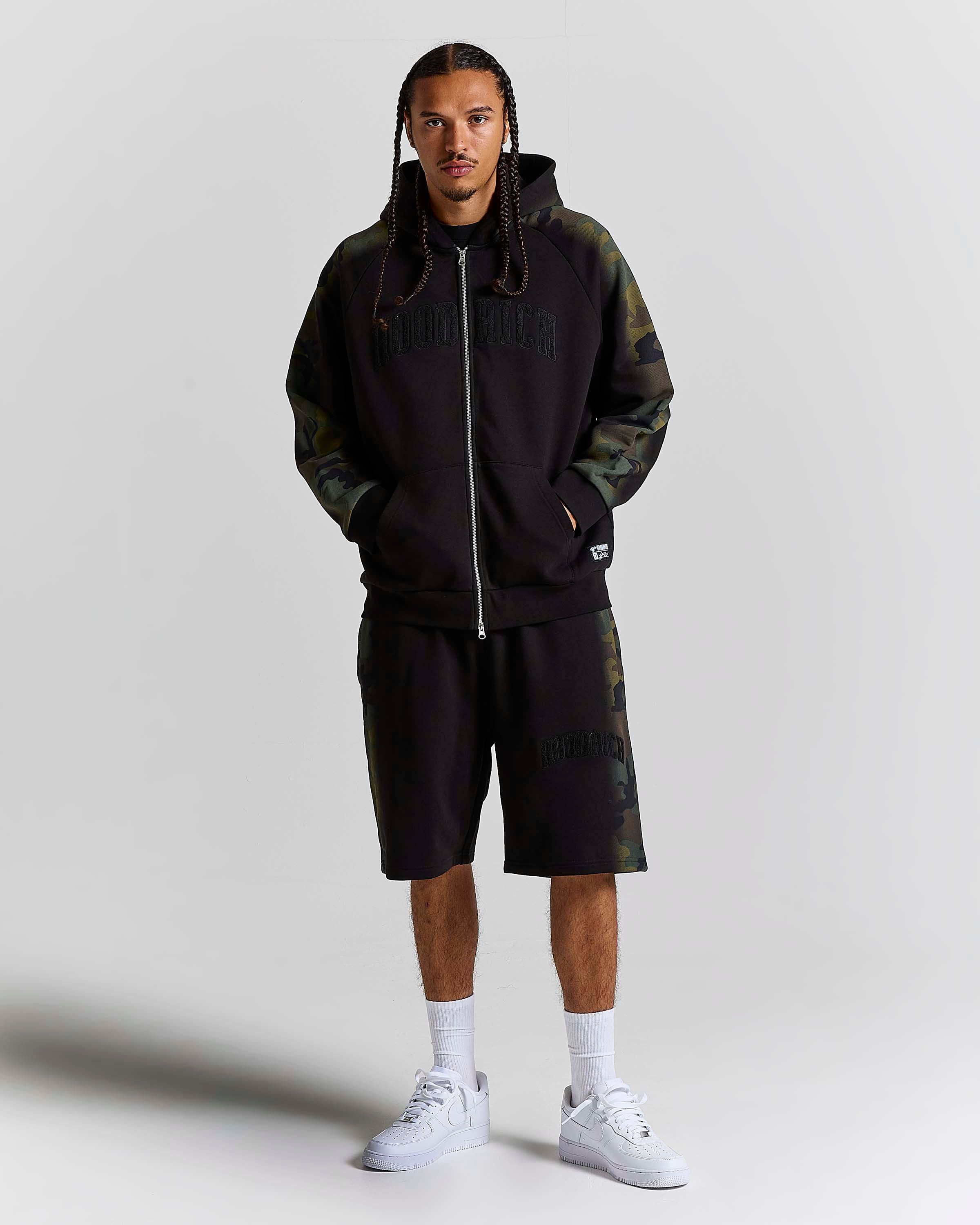 A man modeling a black zip-up hoodie and matching shorts set from HOODRICH, featuring dark green camouflage sleeves and side panels, styled with white sneakers and socks against a light gray studio background.