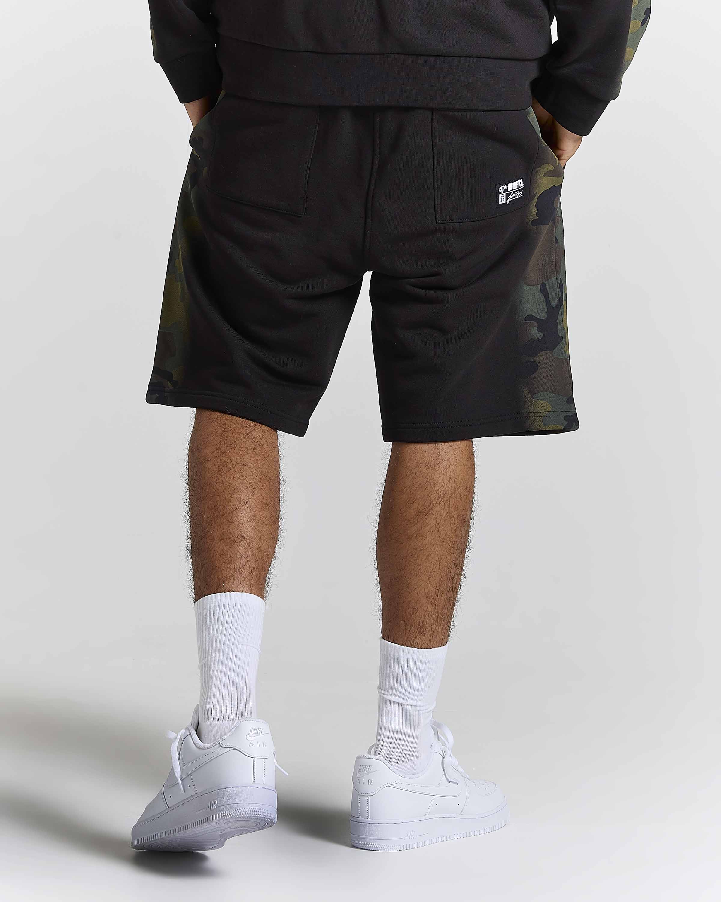 Rear view of a person wearing black athletic shorts with green and brown camouflage side panels, white crew socks, and white low-top sneakers against a light gray studio background.