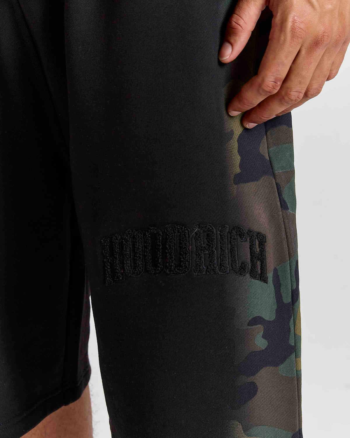 Close-up of a person wearing black sweatpants with a tonal black embroidered 'hoodrich' logo and a green camouflage pattern along the side, against a light gray background.