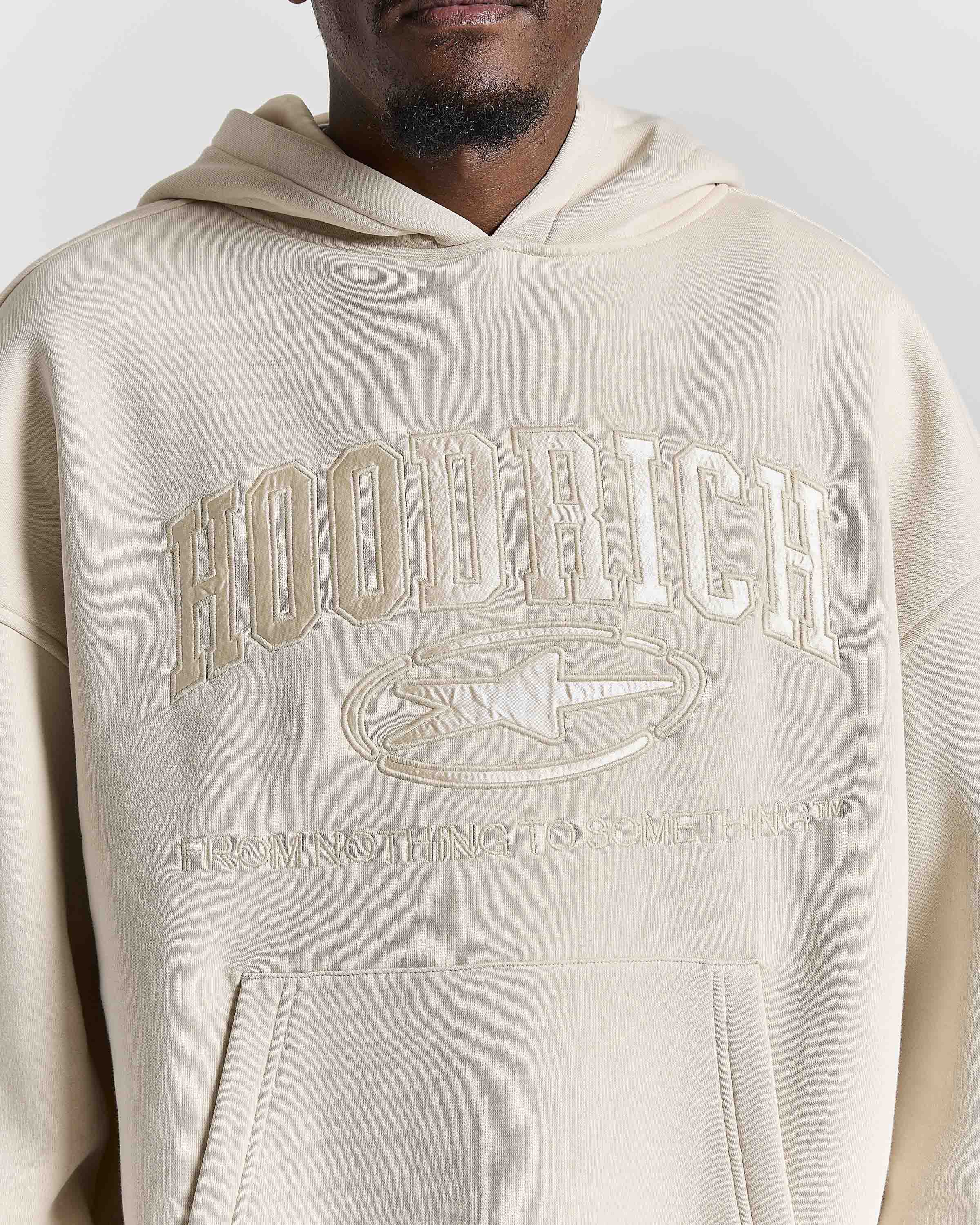 A close-up of a person wearing a cream-colored hoodie with tonal embroidered text reading 'HOODRICH' and 'FROM NOTHING TO SOMETHING' on the front.