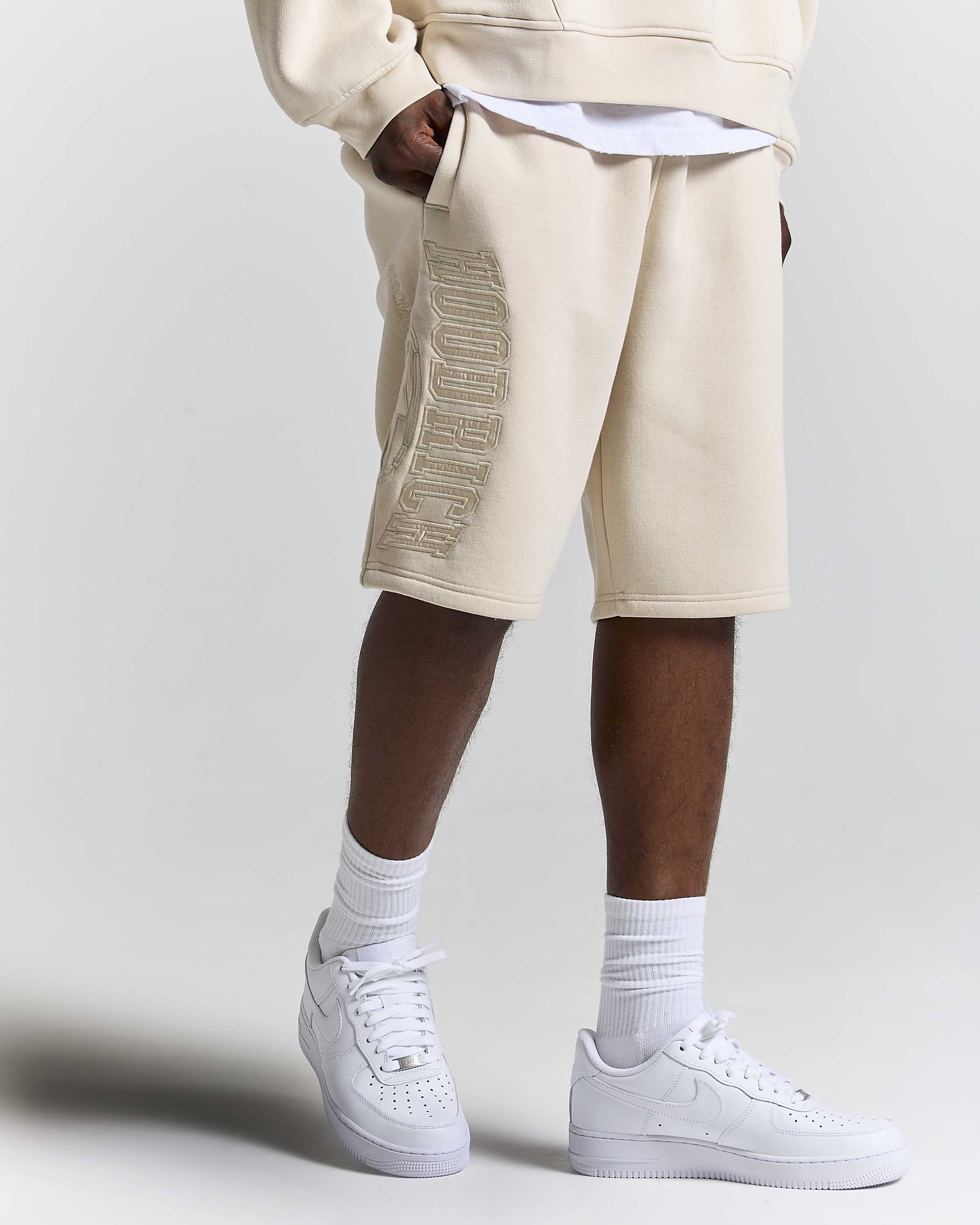 A person wearing beige sweat shorts with a metallic gold embroidered logo down the side, styled with a white t-shirt, white crew socks, and white Nike Air Force 1 sneakers against a light gray studio background.