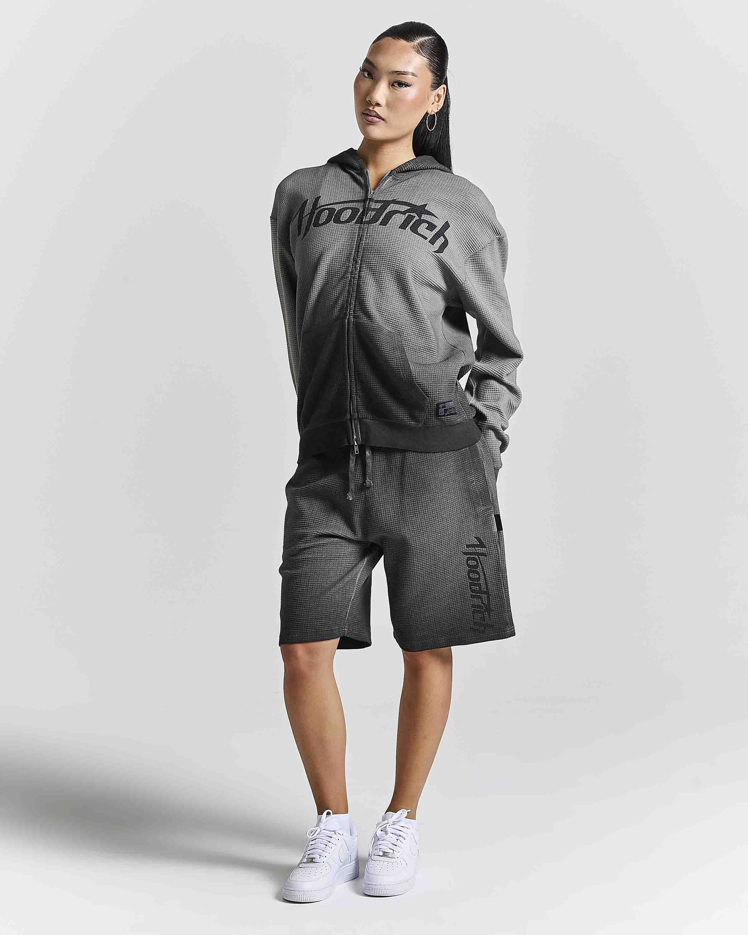 A woman modeling a dark gray two-piece tracksuit featuring a zip-up hoodie with 'HOODRICH' printed across the chest and matching shorts with 'HOODRich' printed down the leg, paired with white sneakers against a light gray studio background.