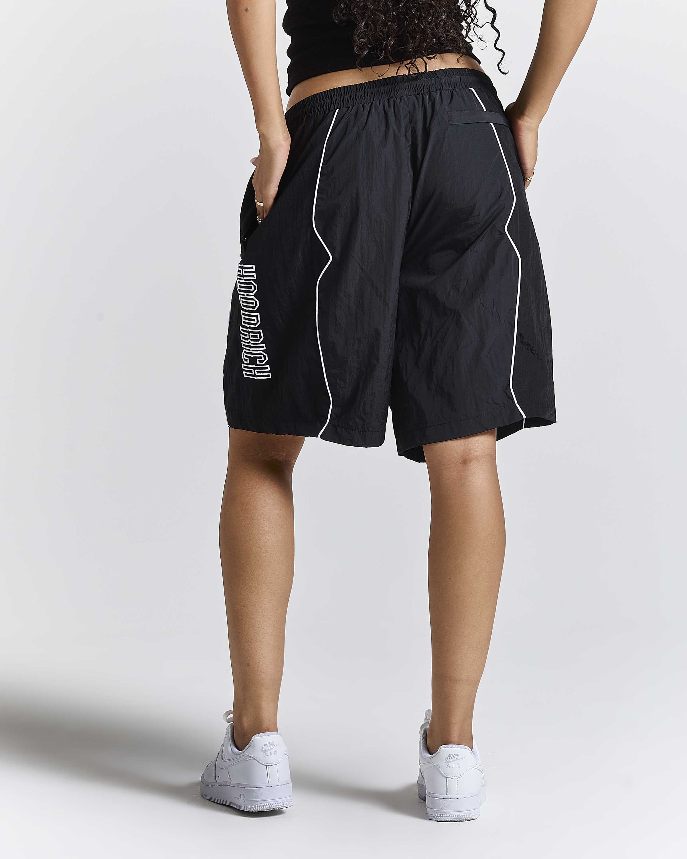 A person shown from the back wearing black athletic shorts with white piping and white sneakers, photographed against a light gray studio background.