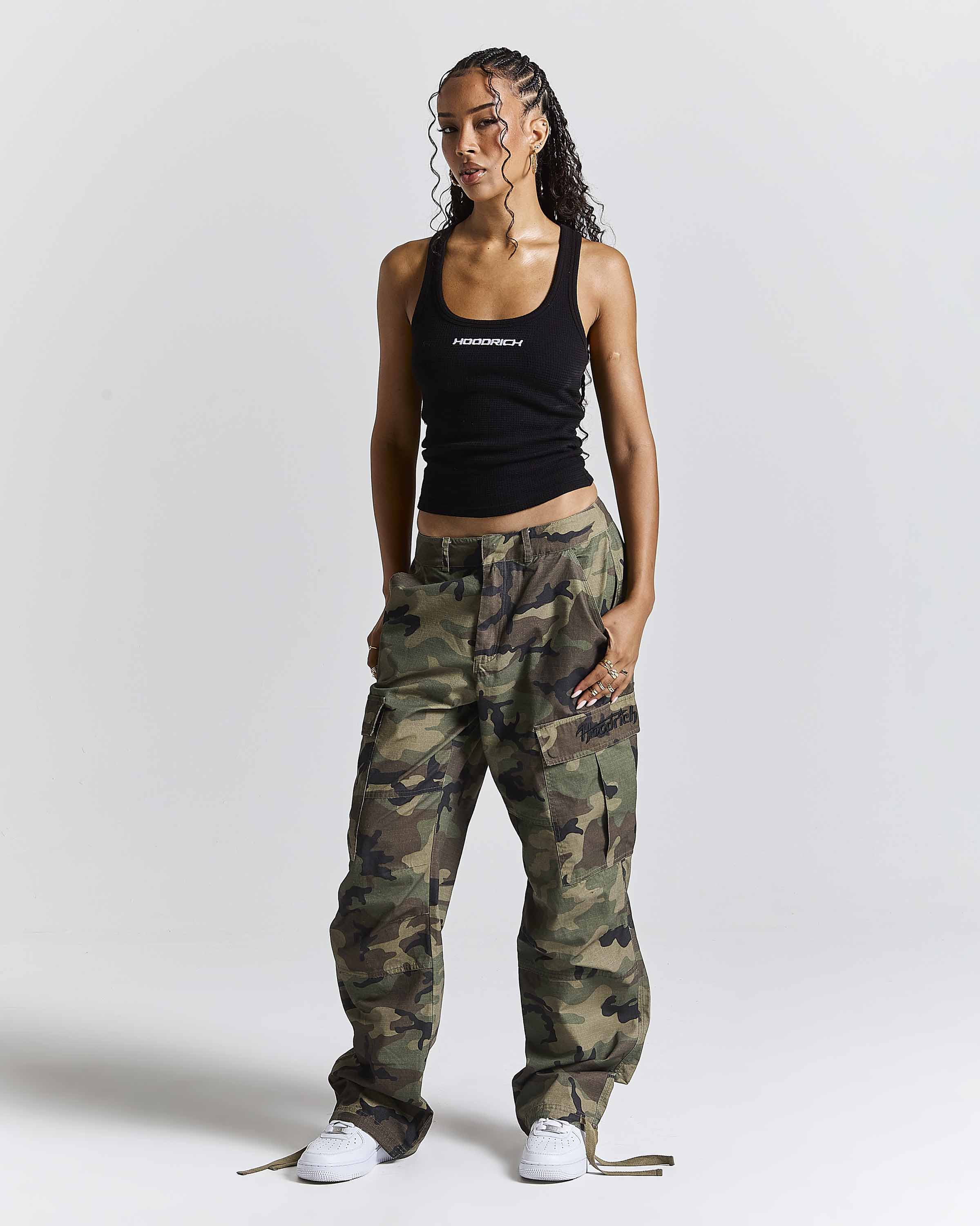 A model stands against a light gray studio background wearing a black tank top with white text, loose camouflage cargo pants, and white sneakers. The tank top has the brand name 'HOODRICH' printed on the chest.