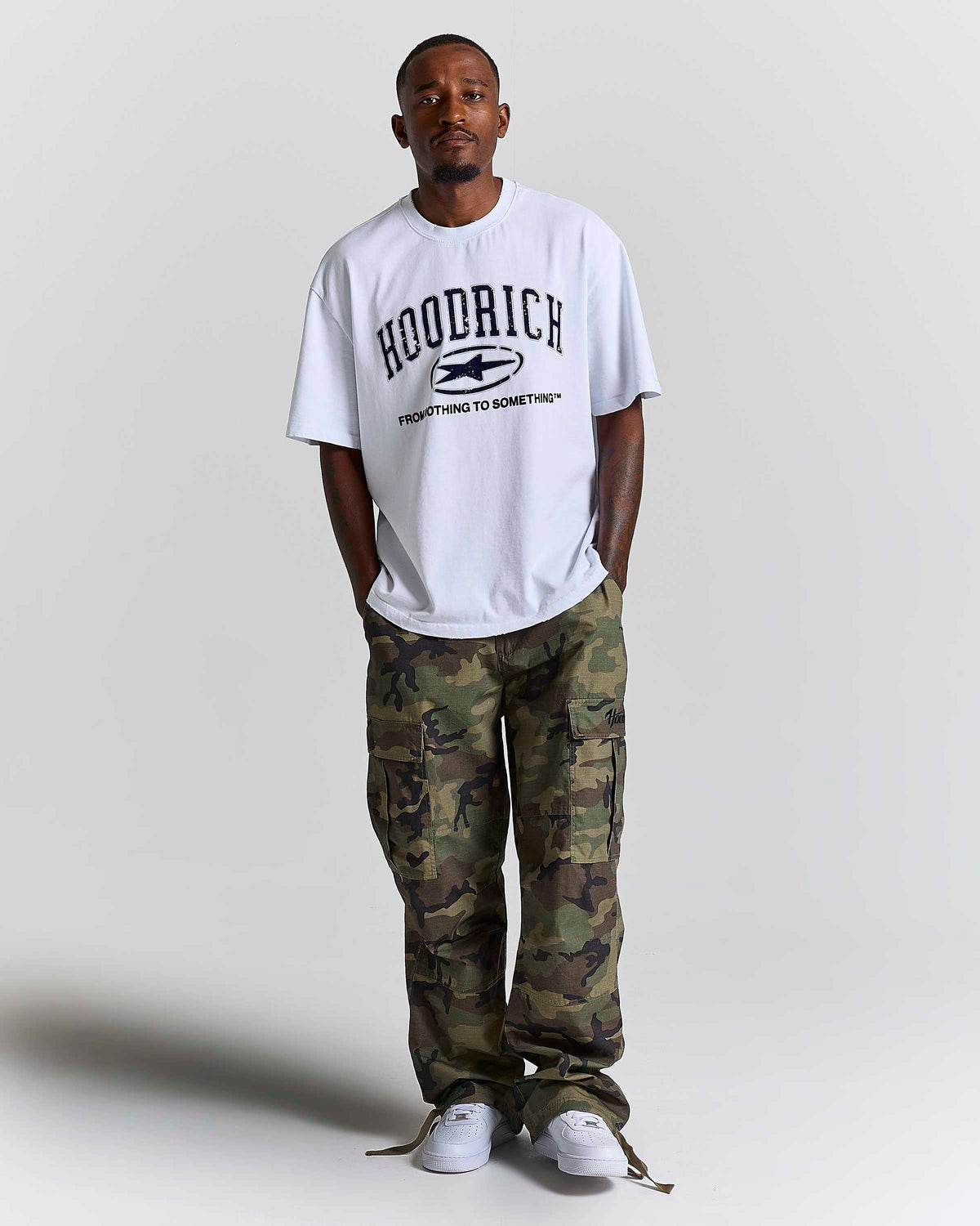 A man wearing a white HOODRICH t-shirt with a black graphic print, paired with green and brown camouflage cargo pants and white sneakers, standing against a light gray studio background.