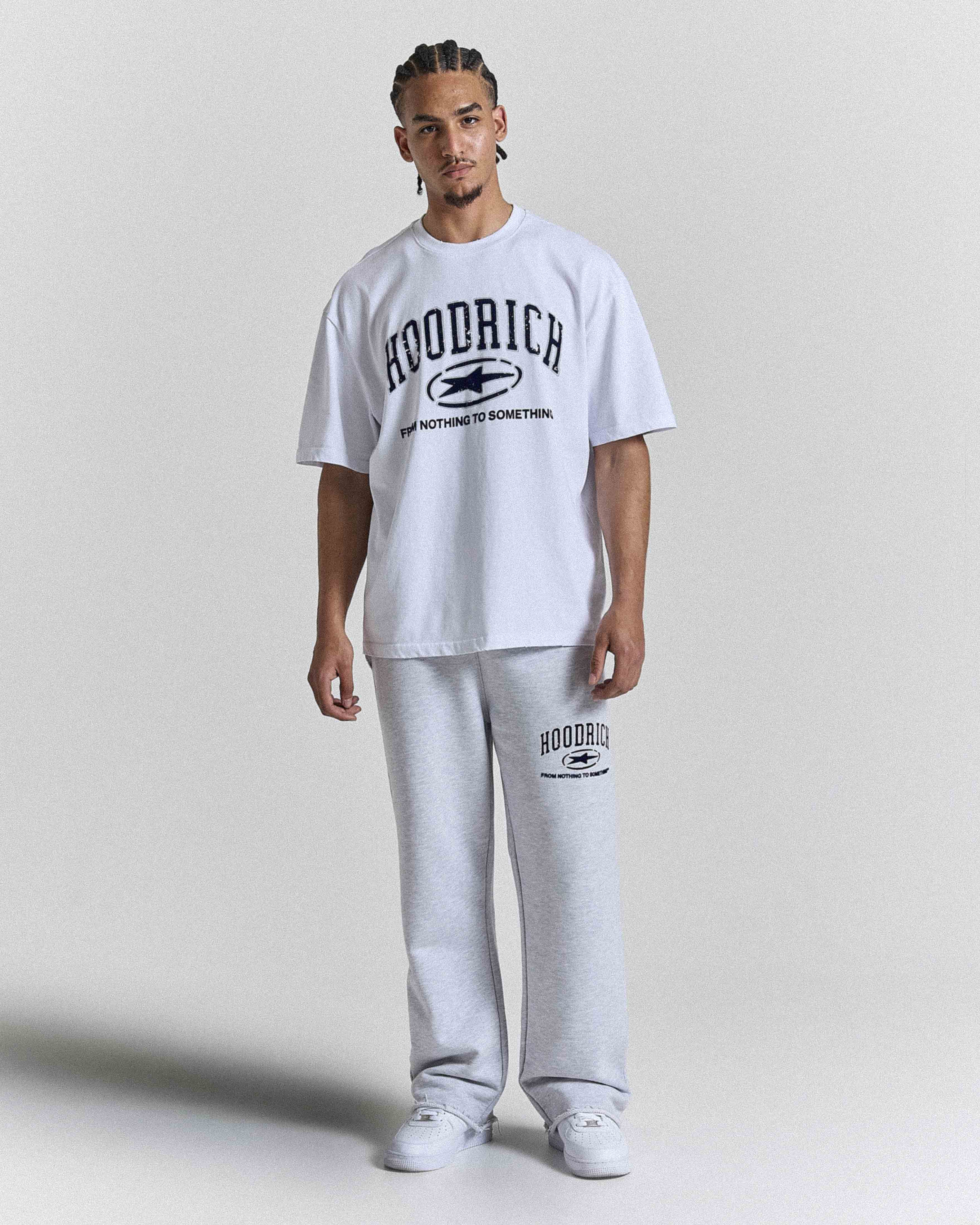 A man modeling a white short-sleeve t-shirt and light gray sweatpants set from the brand Hoodrich, featuring black collegiate-style graphic text and logos on both pieces, against a plain light gray studio background.