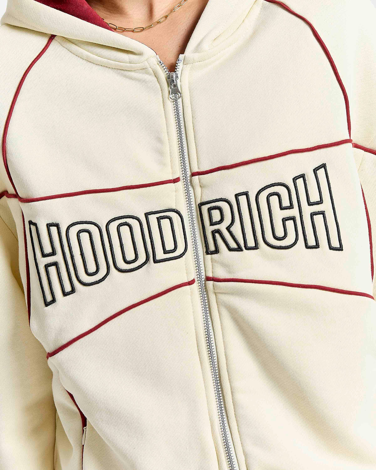 Close-up of a cream zip-up hoodie with 'HOODRICH' embroidered across the chest in black outlined letters, accented by dark red piping lines.
