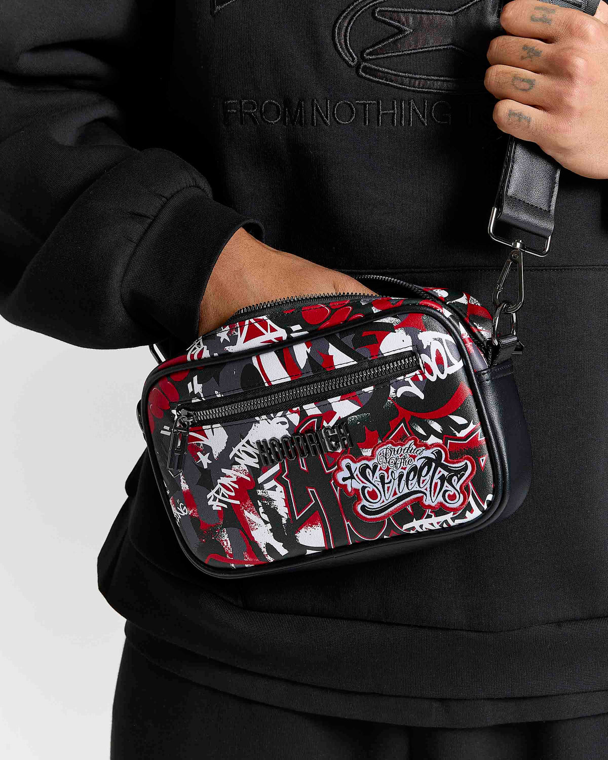 A black crossbody bag with a red and white graffiti pattern, worn by a person in a black sweatshirt. The bag features a top zipper, a front zippered pocket, and a black strap.