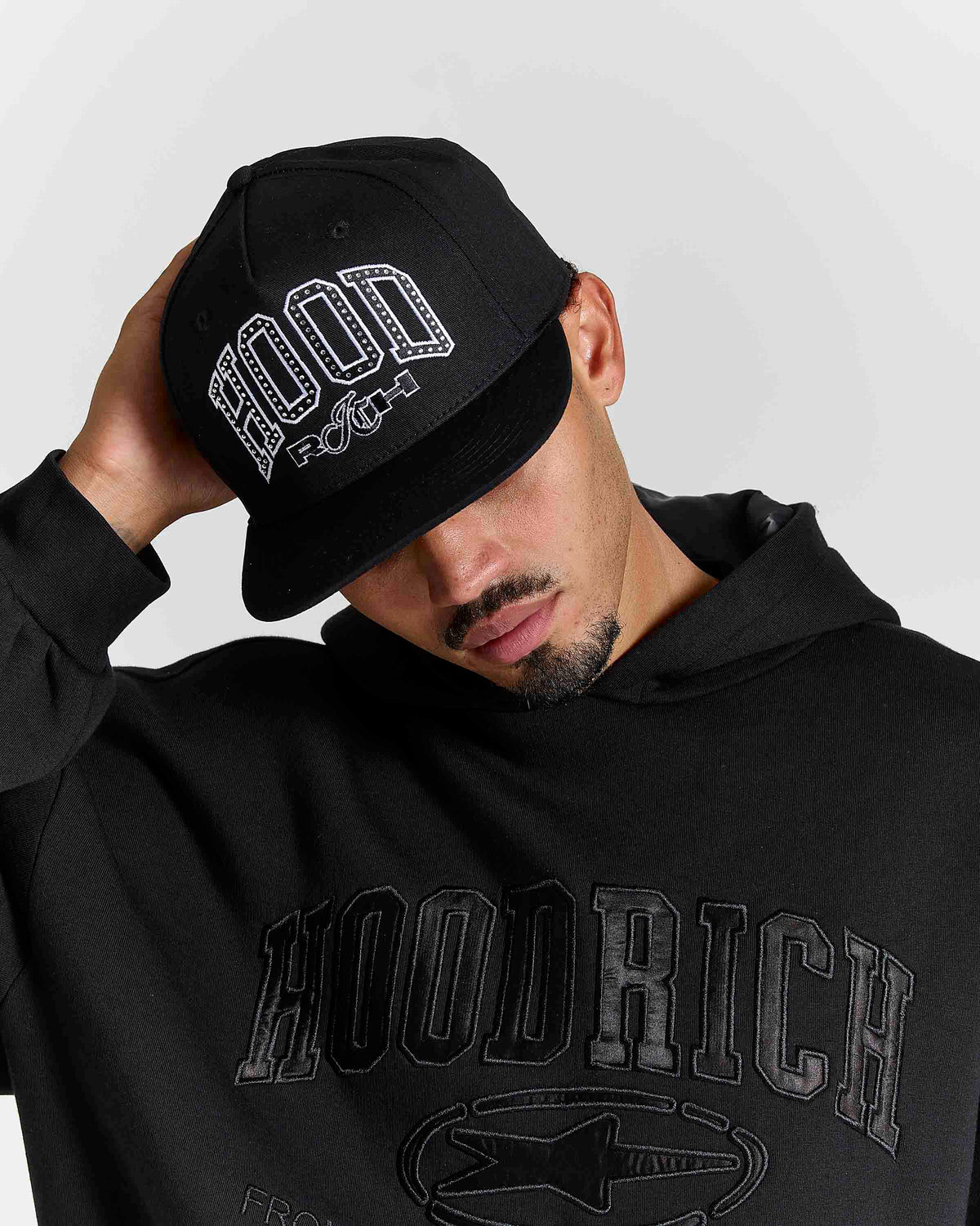 A man wearing a black baseball cap with white 'HOODRICH' text, paired with a black hoodie featuring a tonal 'HOODRICH' graphic on the front, against a light gray background.