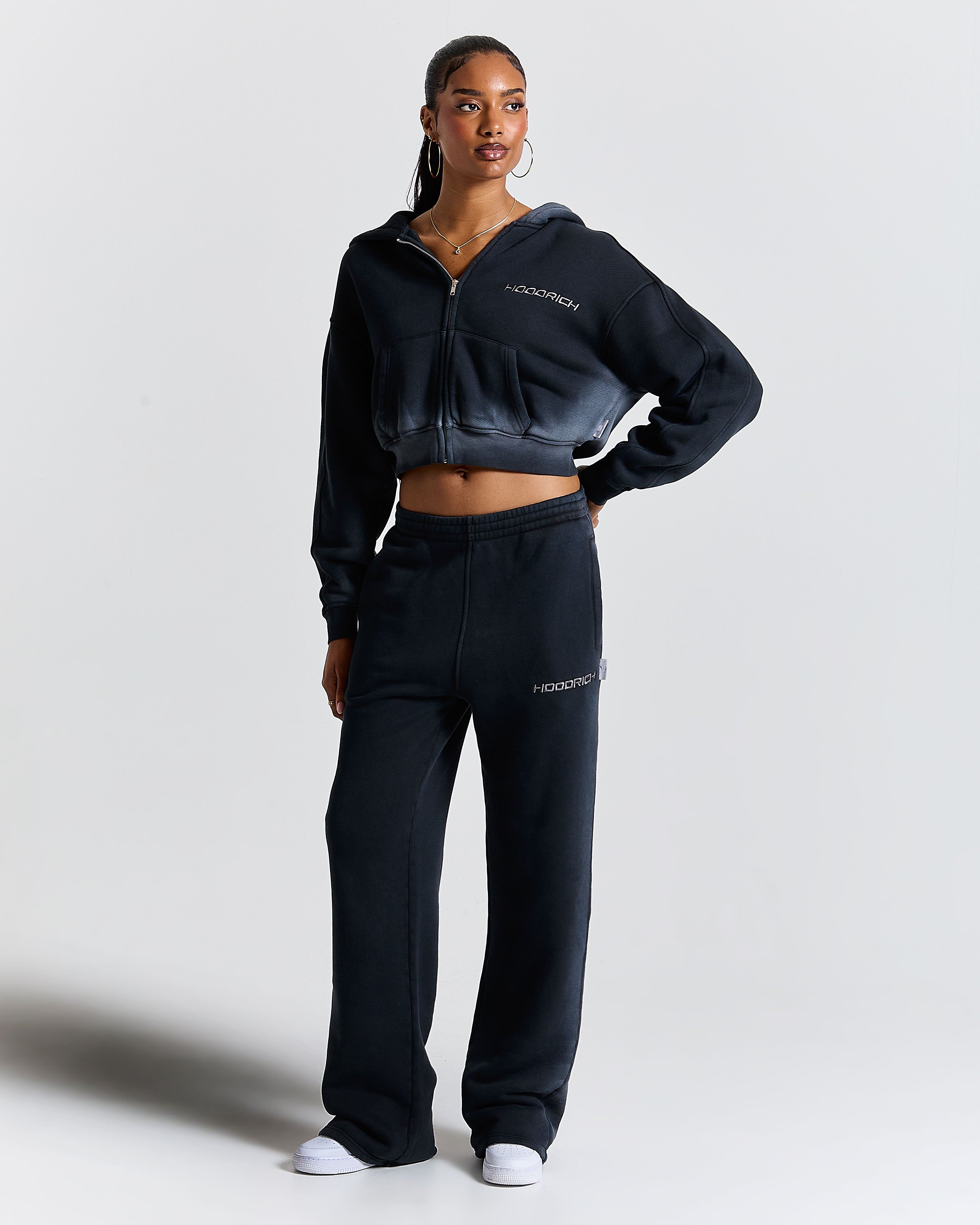 Female model wearing black wide-leg joggers with an embroidered Hoodrich logo on the leg.