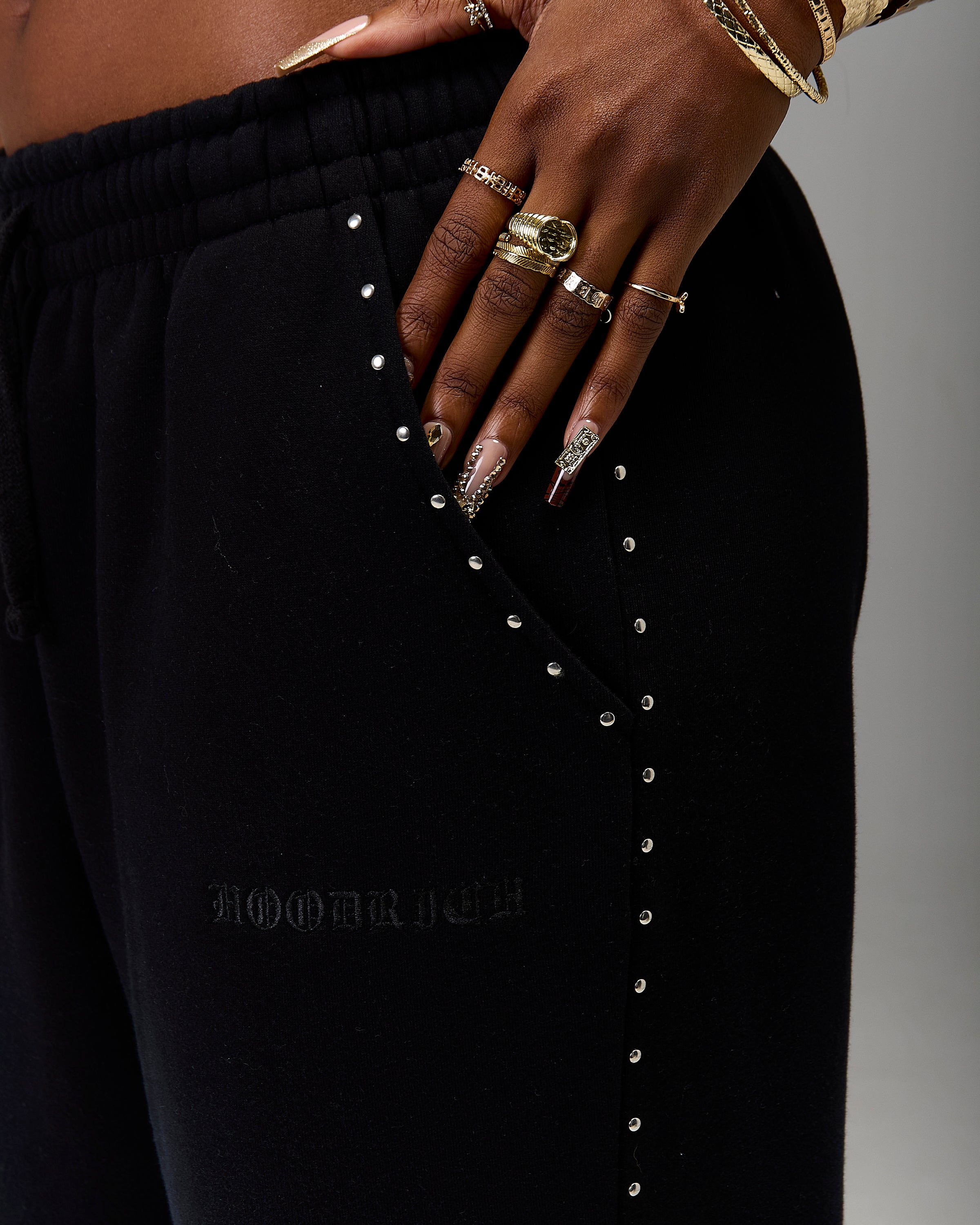 Close-up of a person wearing black jogger sweatpants with silver stud embellishments down the pocket and a subtle embossed logo on the thigh. The person's hand and decorated nails are resting on the waistband, with gold rings and bracelets visible against a neutral gray background.