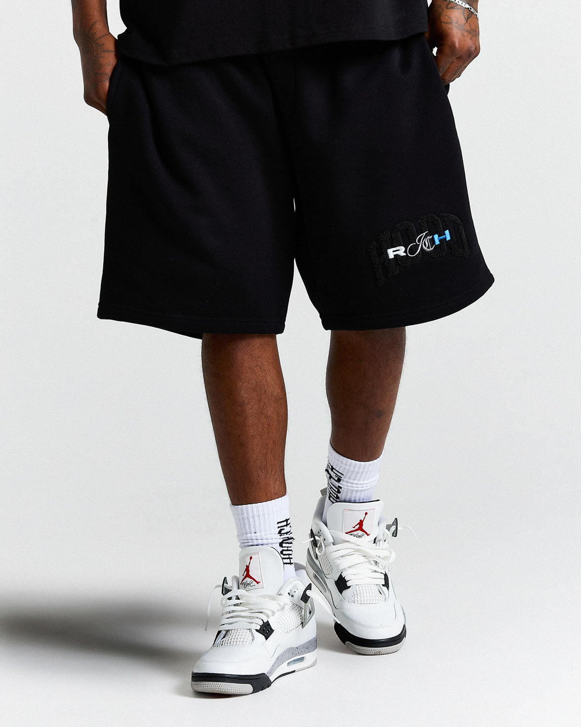 Person wearing black shorts with a logo and white sneakers on a light gray background
