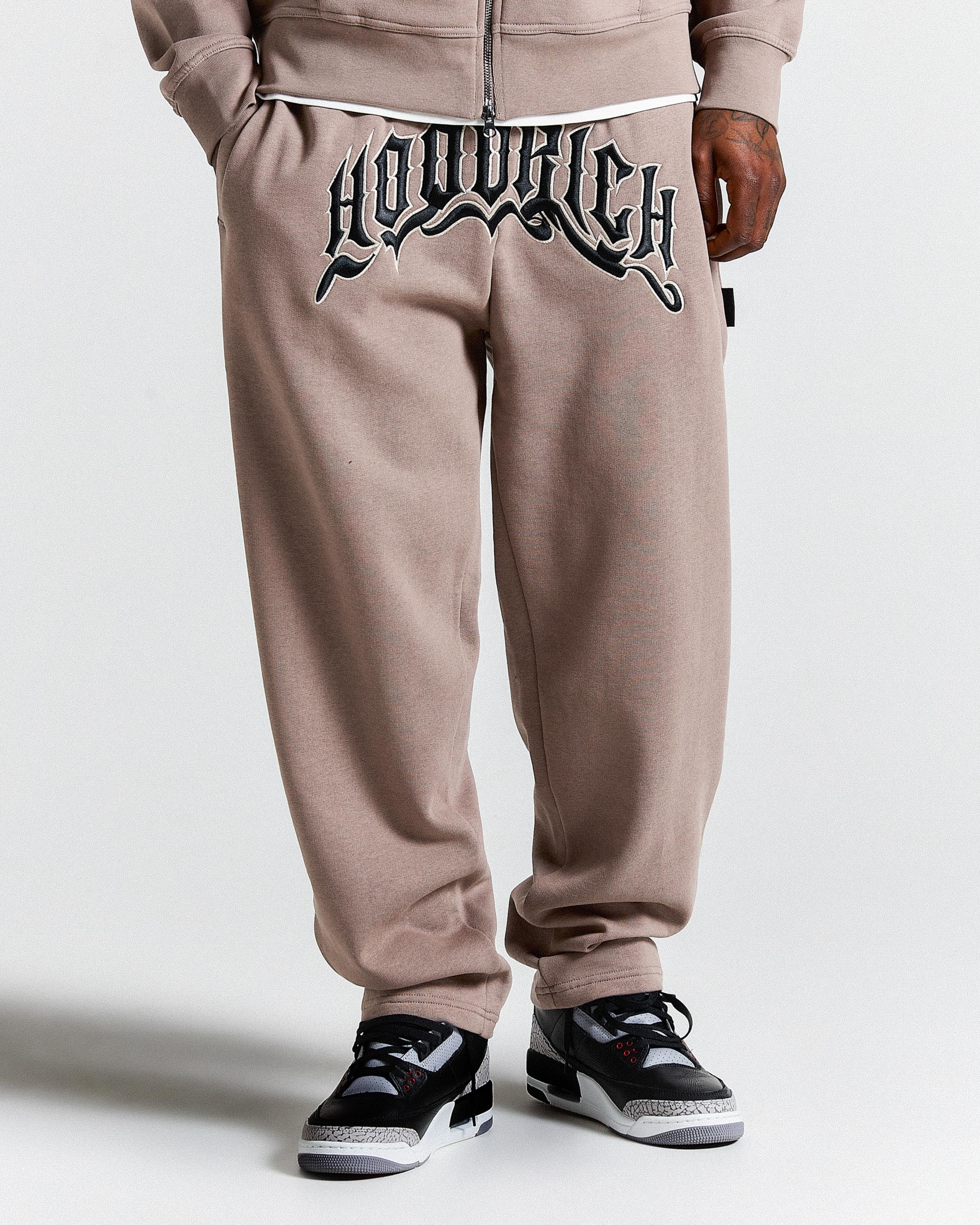 Beige sweatpants with a brand logo on the back, worn by a person, on a white background.