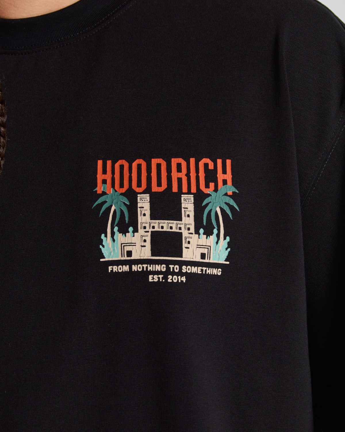 Close-up of a black crewneck sweatshirt featuring a graphic print on the chest with the word 'HOODRICH' in orange, a white gate illustration with teal palm trees, and the text 'FROM NOTHING TO SOMETHING EST. 2014'.