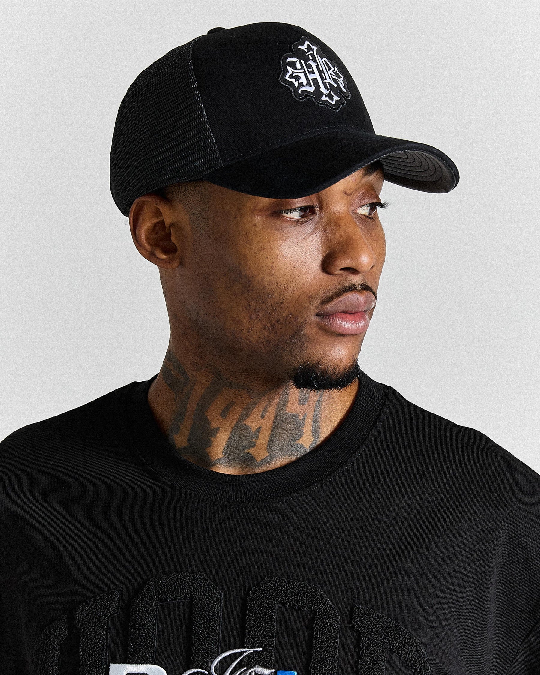 Man wearing a black cap and black shirt with visible tattoos on a gray background