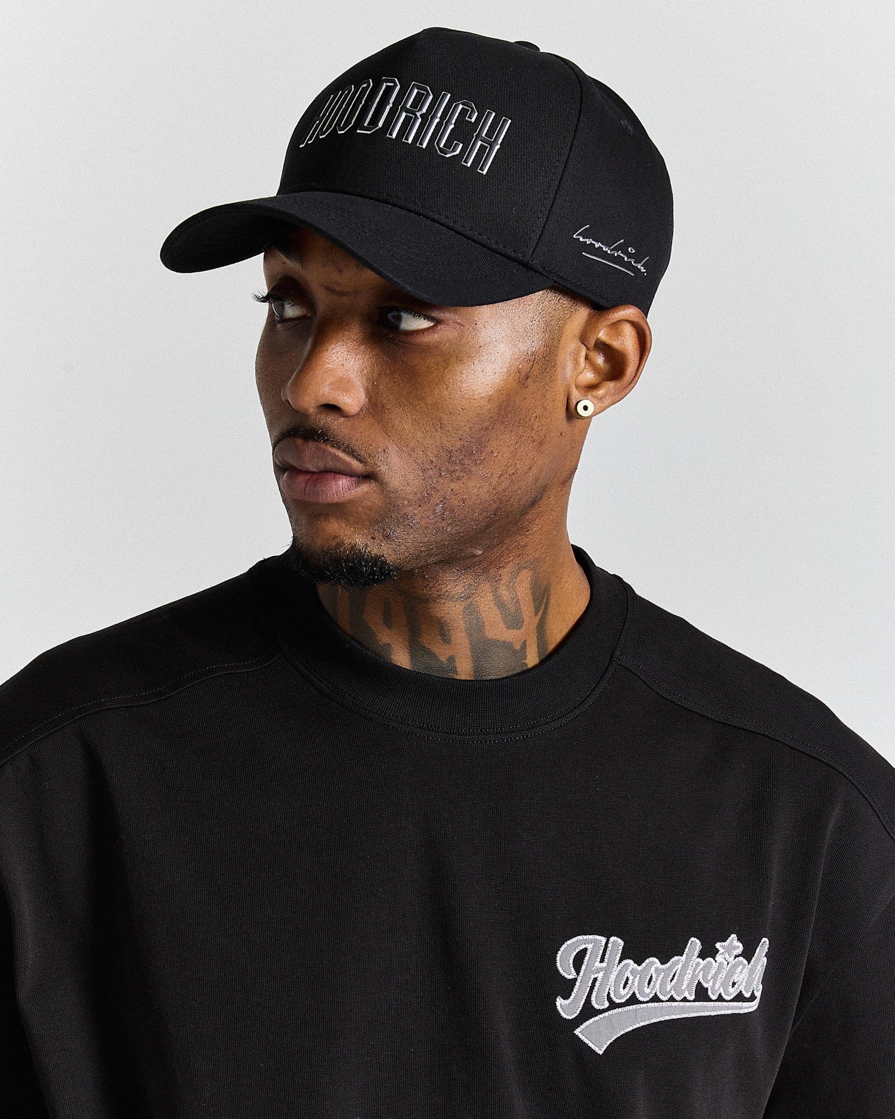Man wearing a black 'Hoodrich' cap and sweatshirt against a gray background