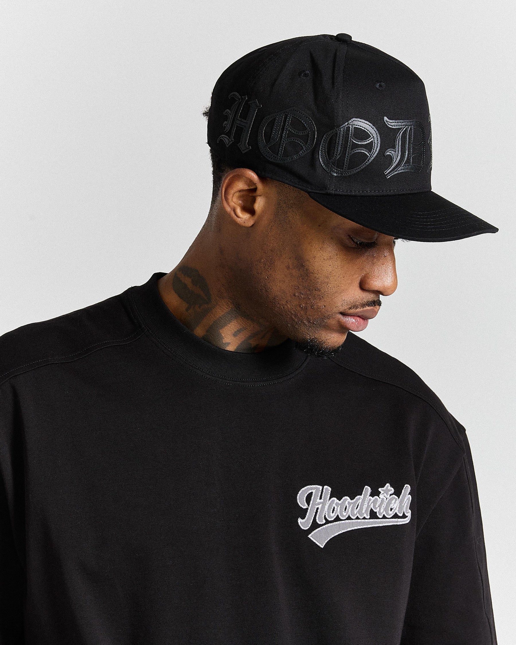 Man wearing a black cap and black hoodie with 'Hoodrich' logo on a gray background