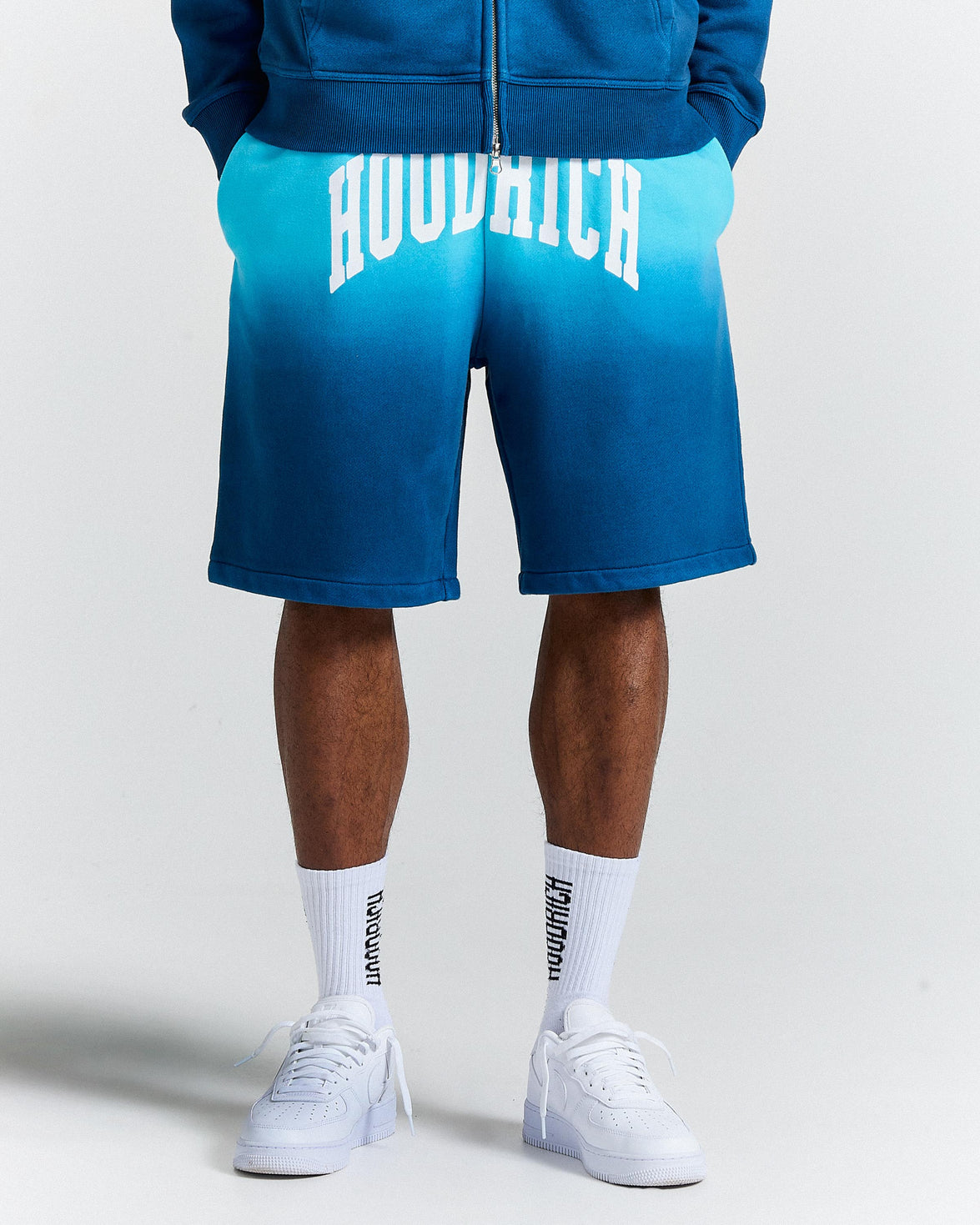 Person wearing blue shorts with 'Hoodrich' branding, white sneakers, and socks on a light gray background