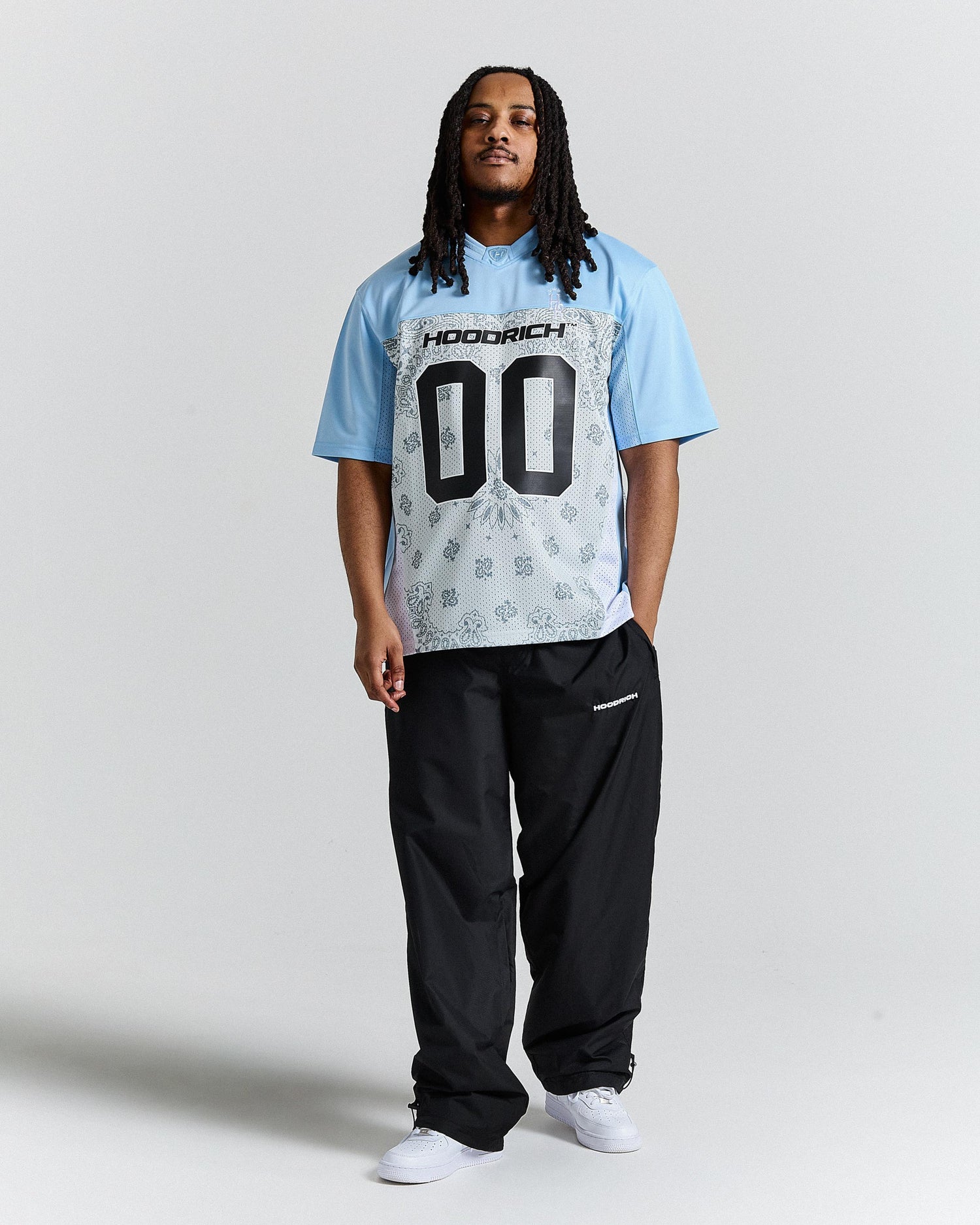 Person wearing a light blue t-shirt with text and graphics, black pants, and white shoes on a plain background